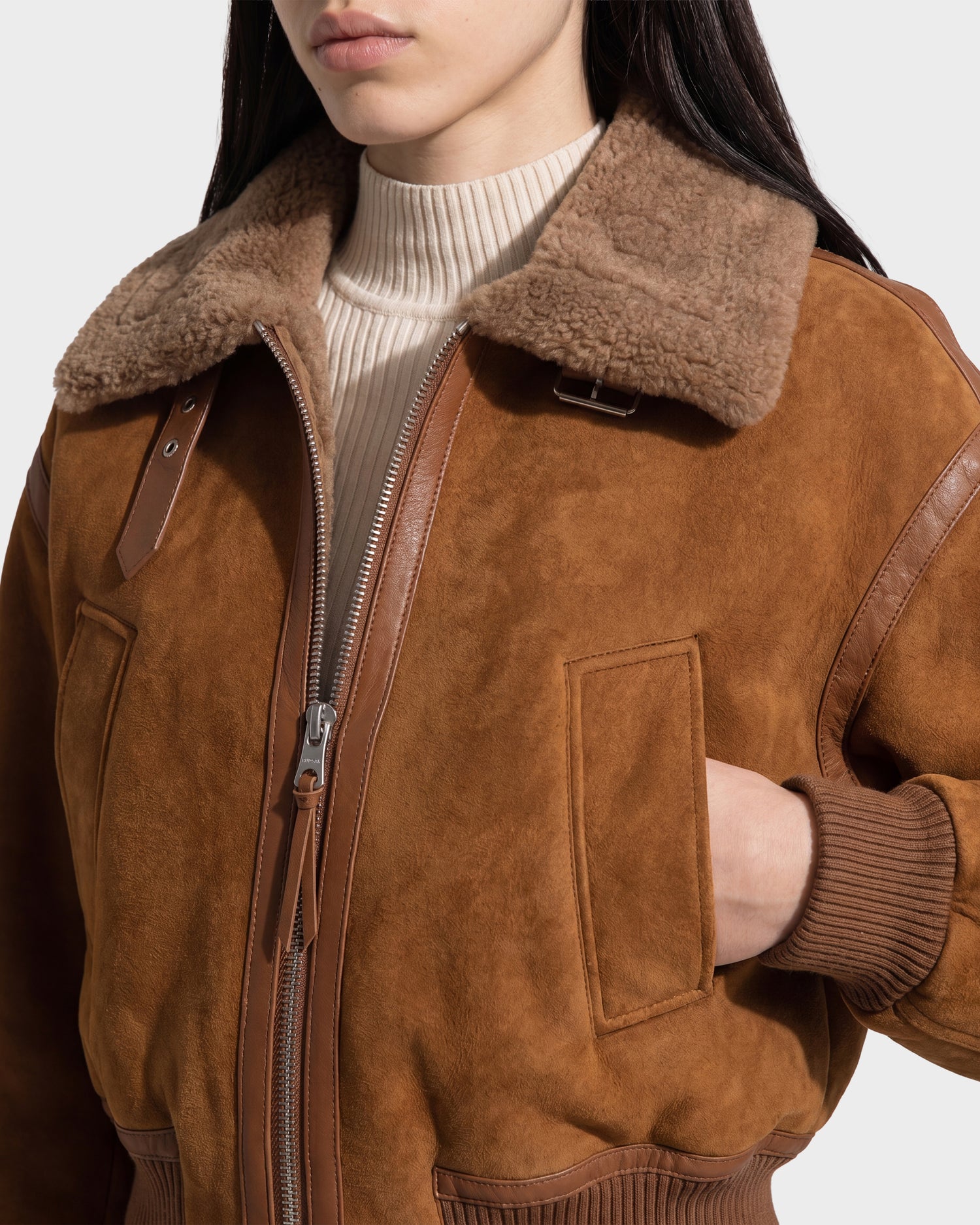 A woman wearing the Rudsak LIZZIE - 8126787 Tan shearling aviator jacket with a wide collar and a tan ribbed turtleneck sweater, her left hand in her pocket.