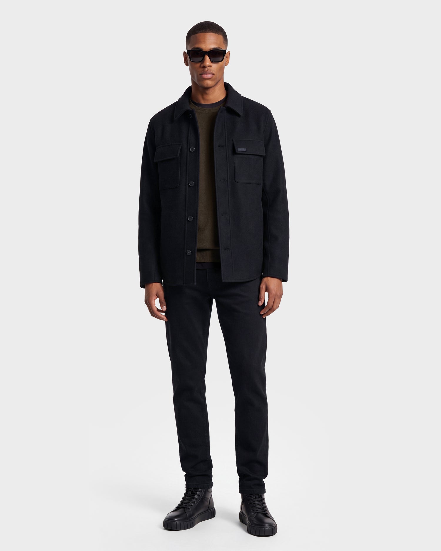 A man wearing sunglasses, black pants, and black sneakers stands against a plain white background, facing forward with a neutral expression while sporting the Rudsak MACKENZIE - 6126715 Black wool blend men’s shirt jacket—an effortless example of transitional dressing.