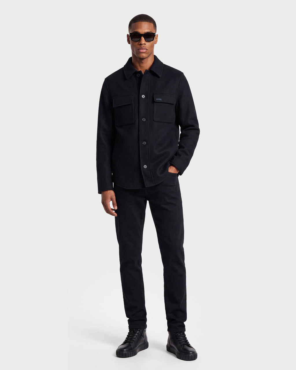 A man stands against a plain background wearing black sunglasses and the Rudsak MACKENZIE - 6126715 Black men’s shirt jacket, crafted from a wool blend and featuring chest pockets—perfect for transitional layering with his black pants and black high-top shoes.