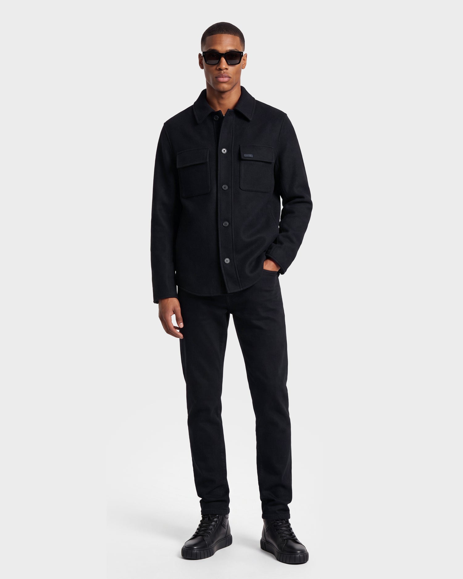 A man stands against a plain background wearing black sunglasses and the Rudsak MACKENZIE - 6126715 Black men’s shirt jacket, crafted from a wool blend and featuring chest pockets—perfect for transitional layering with his black pants and black high-top shoes.