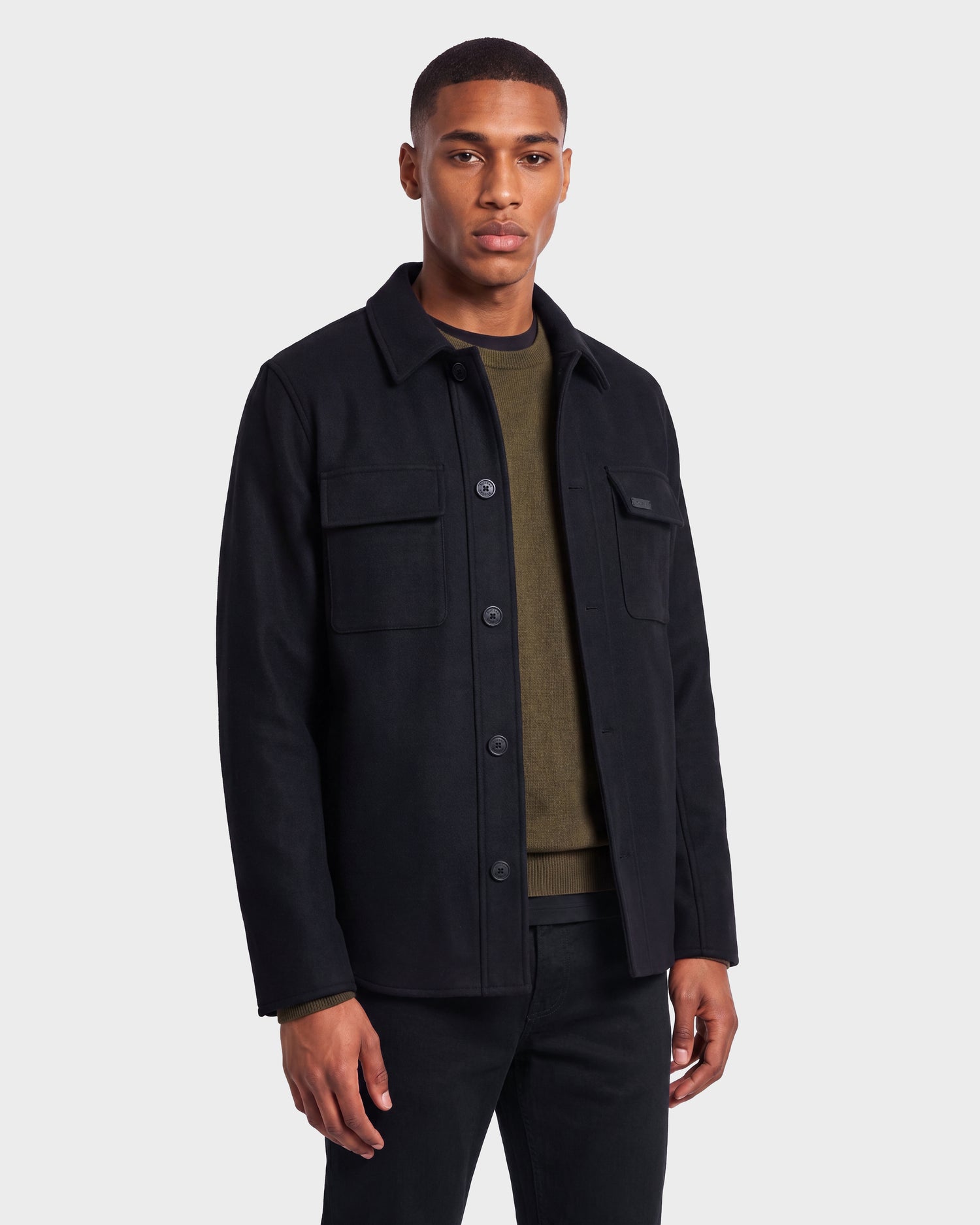 A man stands against a plain background wearing the Rudsak MACKENZIE - 6126715 Black wool blend men’s shirt jacket over an olive green shirt and black pants, facing forward with a neutral expression—an ideal look for effortless transitional dressing.