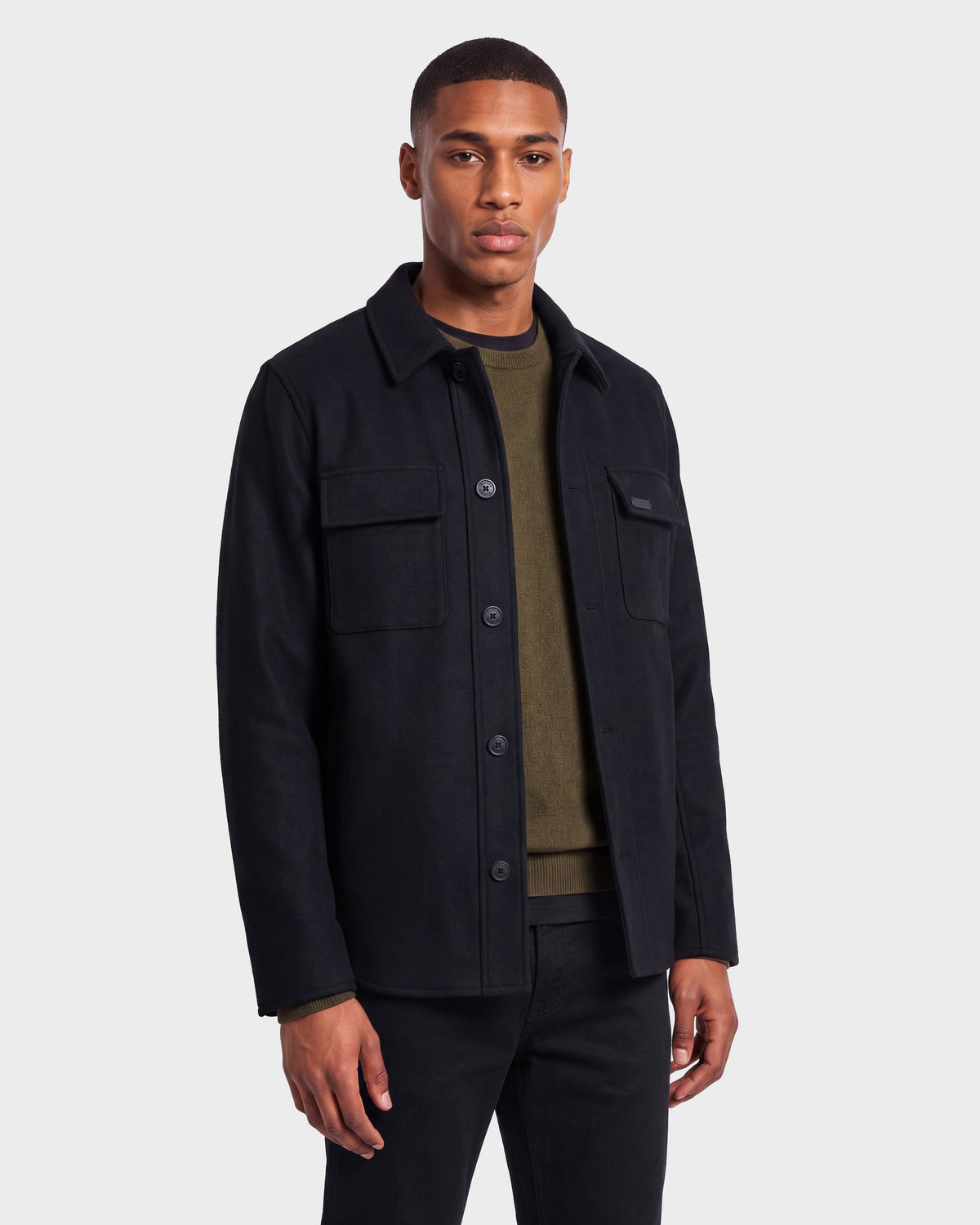 A man stands against a plain background wearing the Rudsak MACKENZIE - 6126715 Black wool blend men’s shirt jacket over an olive green shirt and black pants, facing forward with a neutral expression—an ideal look for effortless transitional dressing.