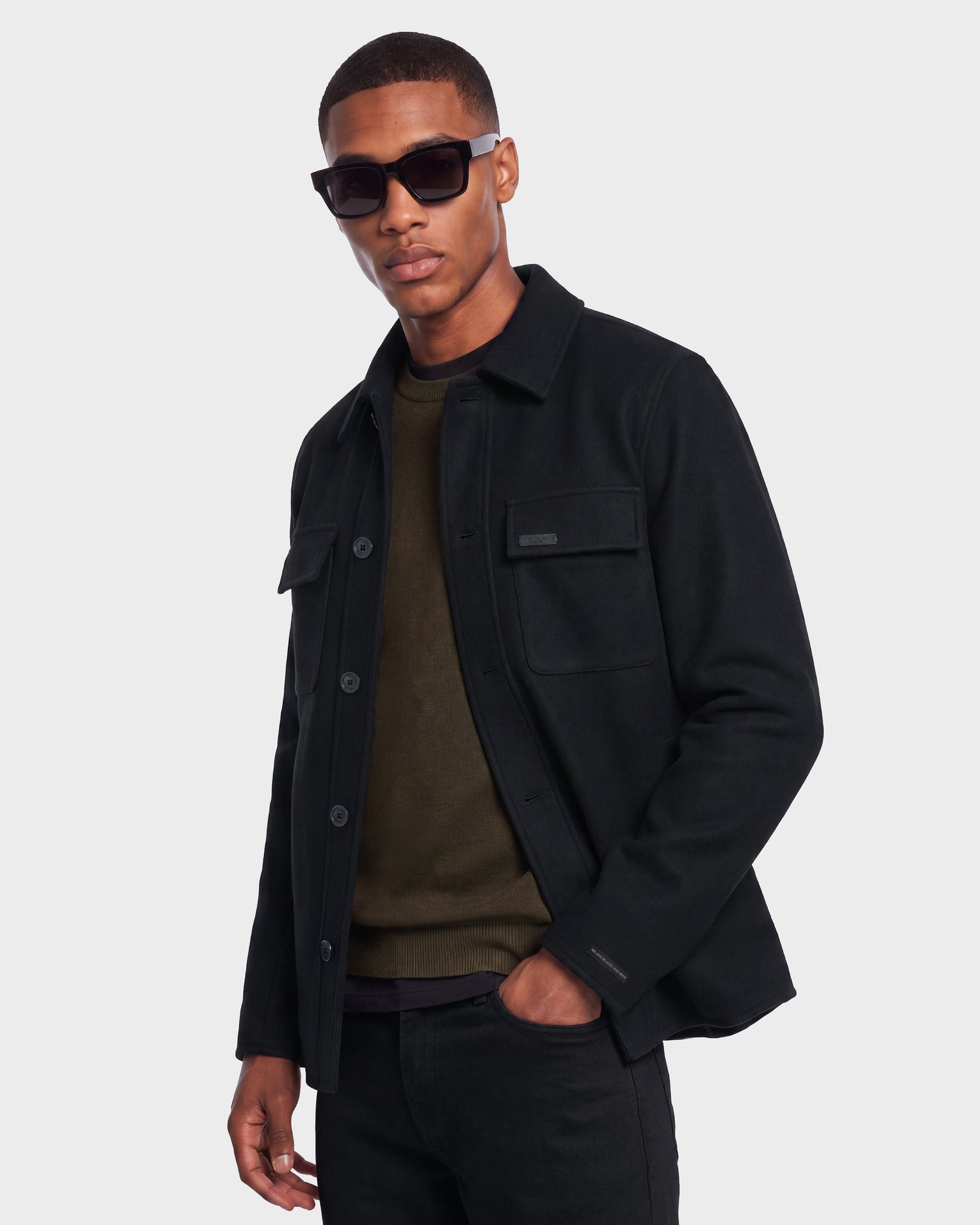 Wearing the Rudsak MACKENZIE - 6126715 Black wool-blend men’s shirt jacket over an olive green sweater, paired with black pants and dark sunglasses, a man stands against a plain light gray background—effortlessly showcasing transitional style.
