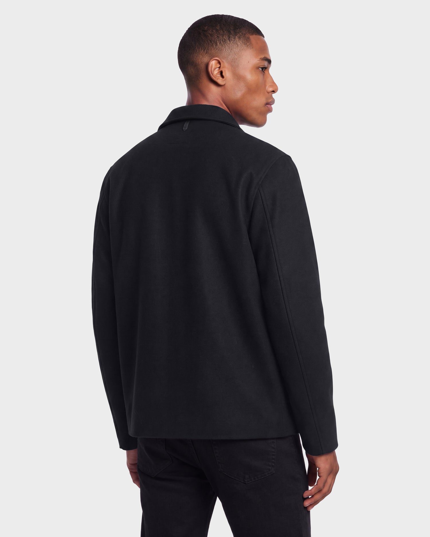 A man stands facing away, wearing the Rudsak MACKENZIE - 6126715 Black wool blend men’s shirt jacket and black pants against a plain, light gray background.
