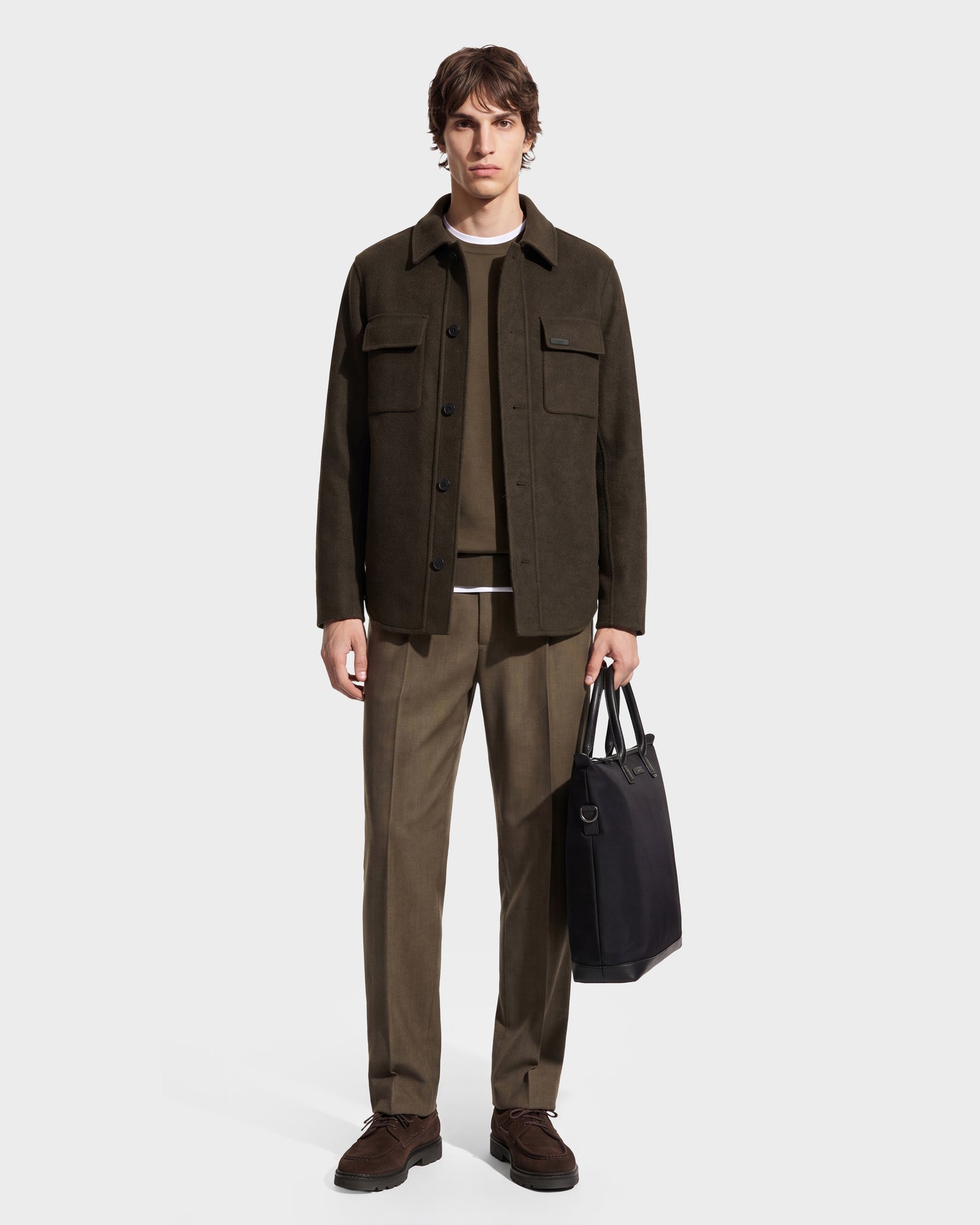 A man wearing the Rudsak MACKENZIE - 6126715 Olive wool blend shirt jacket, taupe pants, and brown shoes stands against a white background, holding a black tote bag in his left hand.