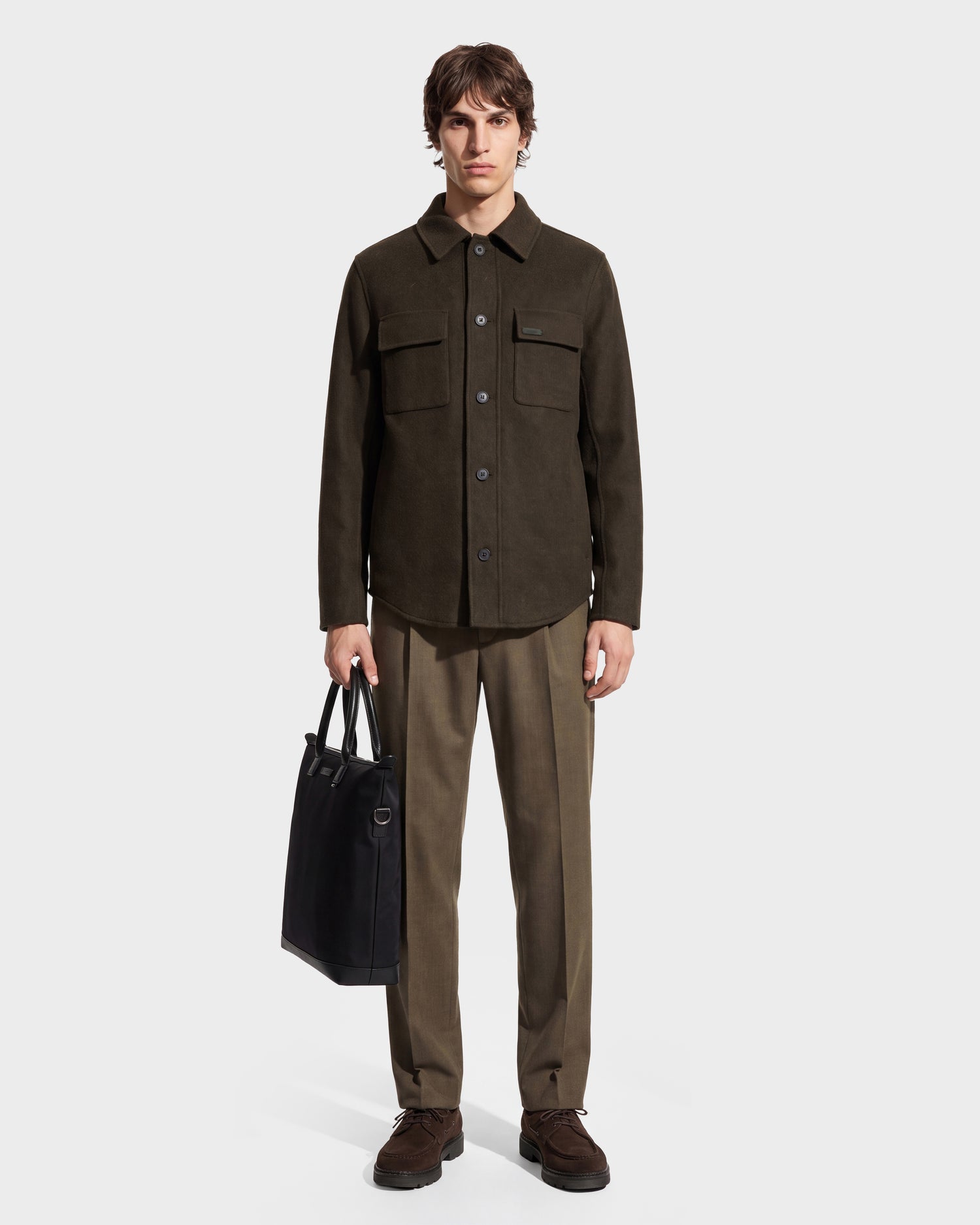 A man stands facing forward against a plain white background, wearing the Rudsak MACKENZIE - 6126715 Olive wool blend men’s shirt jacket with matching trousers and brown shoes, while holding a black tote bag in his left hand.
