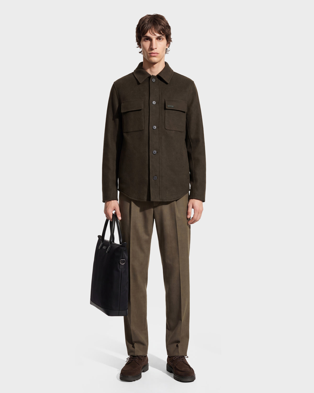 A man stands facing forward against a plain white background, wearing the Rudsak MACKENZIE - 6126715 Olive wool blend men’s shirt jacket with matching trousers and brown shoes, while holding a black tote bag in his left hand.