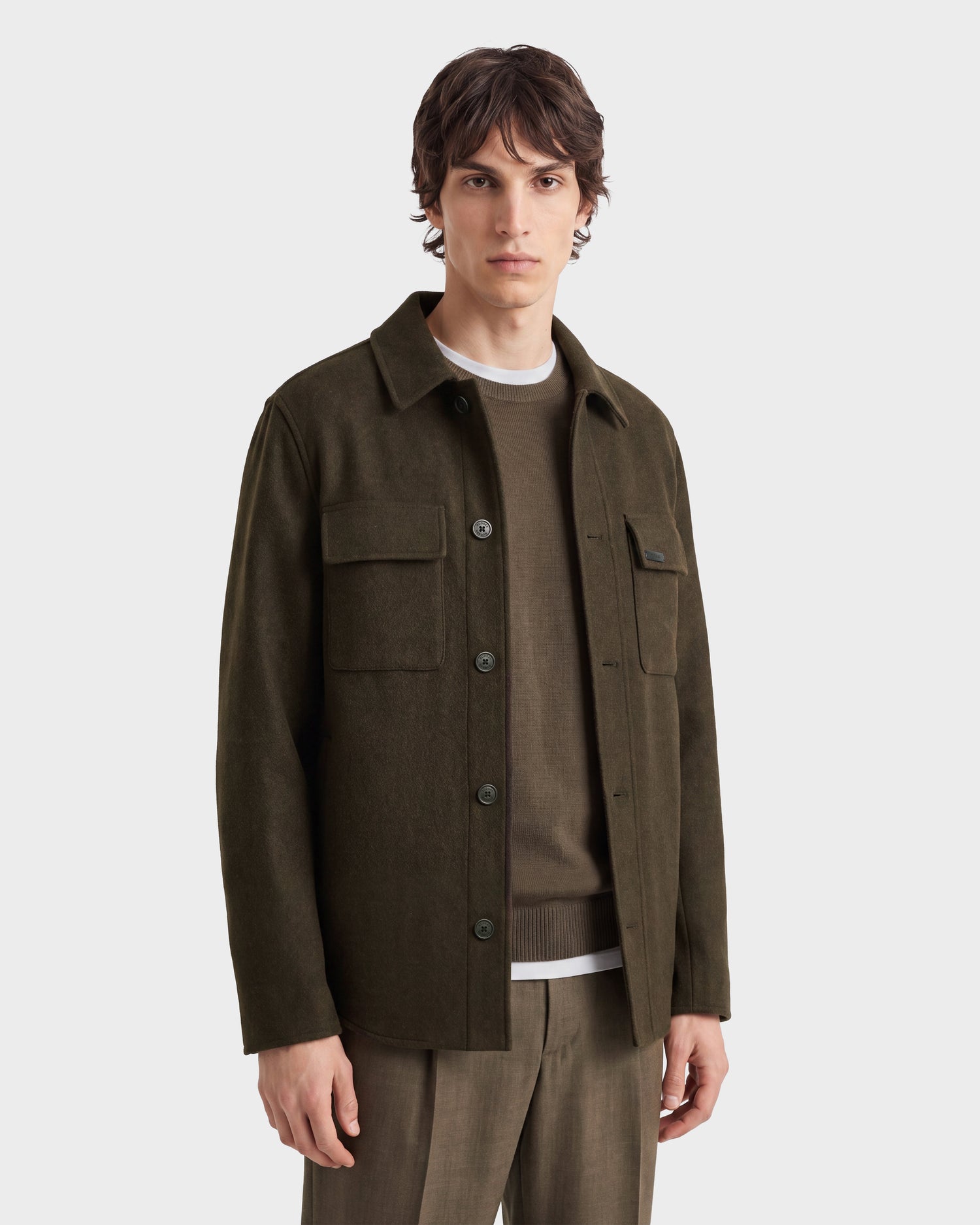A person stands facing forward against a plain light background, wearing the MACKENZIE - 6126715 Olive men’s shirt jacket by Rudsak—a luxurious dark olive green wool blend—layered over a brown sweater and white shirt, paired with brown pants.