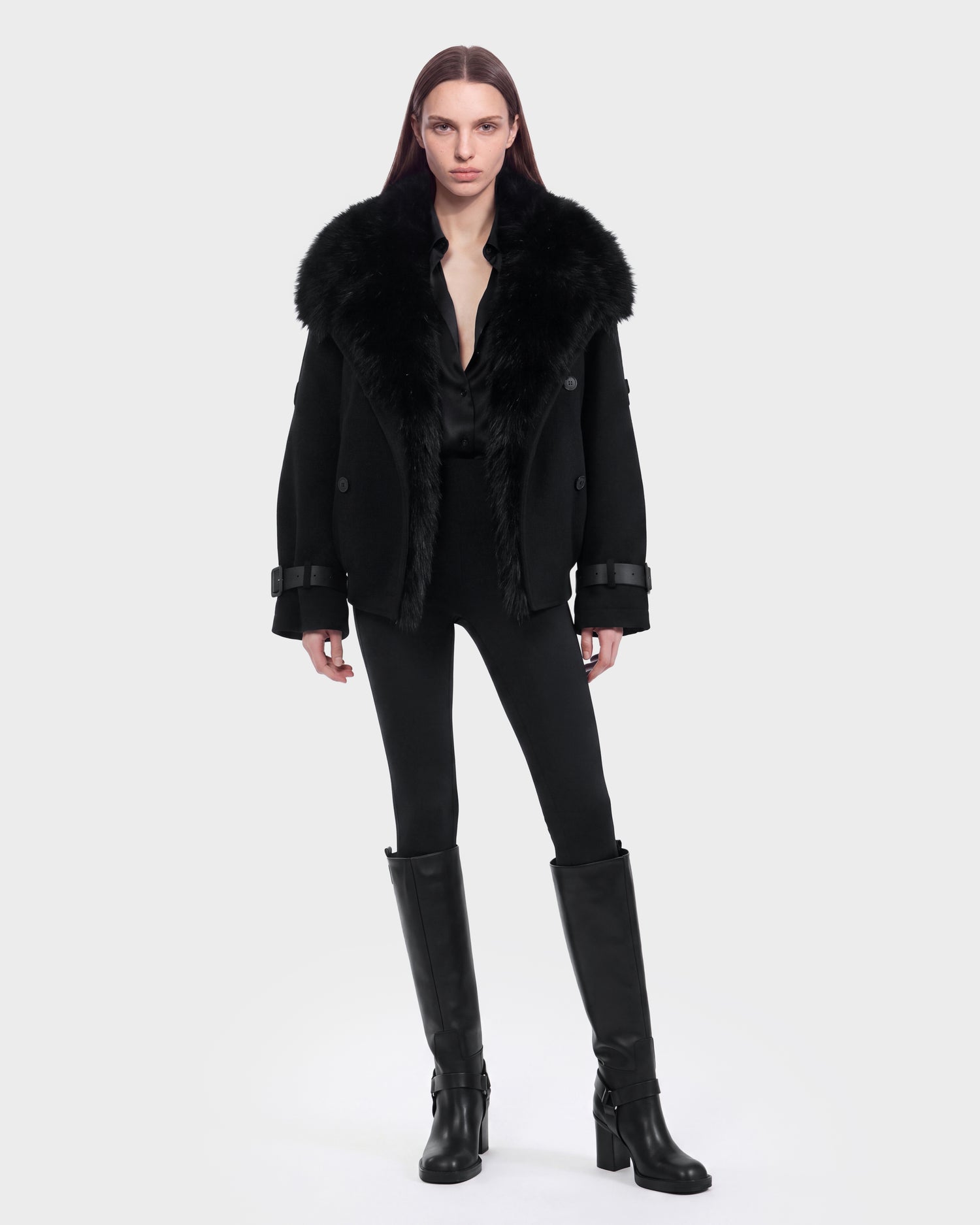 A person with straight, long brown hair wears the Rudsak MAEMI - 8126985 Black fur-collared coat paired with a black shirt, fitted black pants, and knee-high black boots, standing against a plain light gray background. This striking look is available for pre-order now and is expected to ship by September 15th.