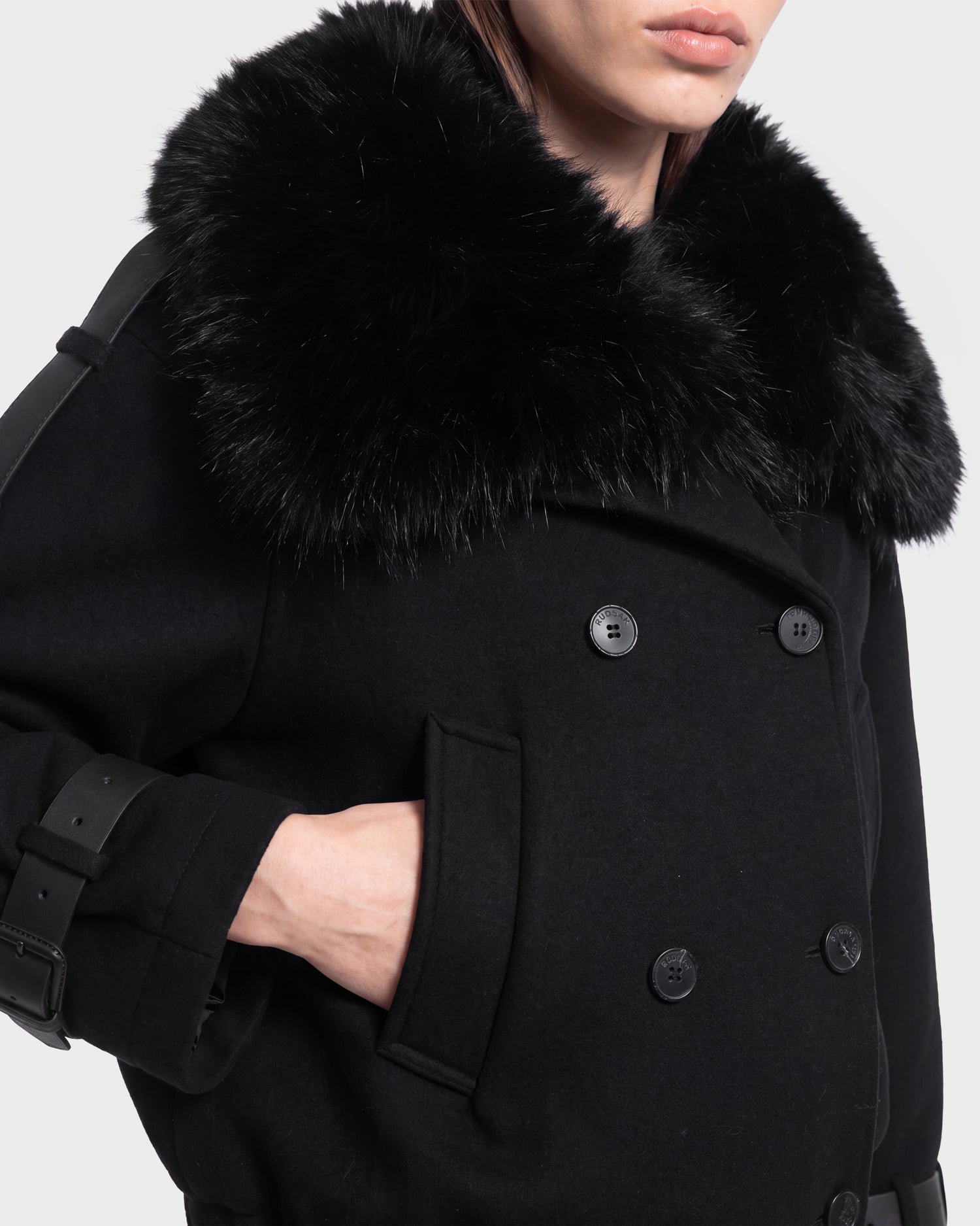 A woman is seen wearing the MAEMI - 8126985 Black coat by Rudsak, featuring a double-breasted design and a luxurious oversized black faux fur collar. Her hand rests in her pocket, and her face is partially visible. This elegant piece is available for pre-order now and is expected to ship by September 15th.