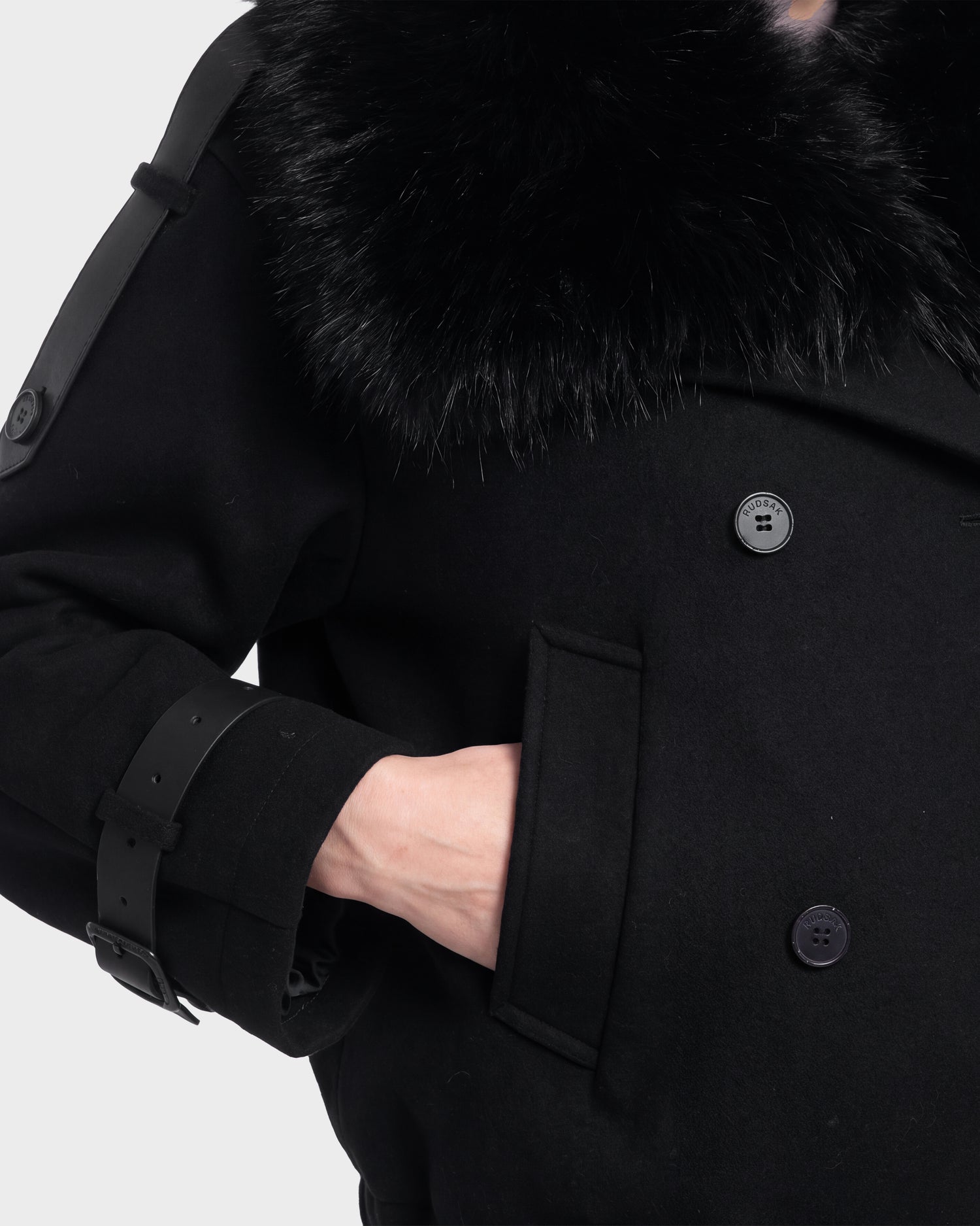 A person wearing the MAEMI - 8126985 Black coat by Rudsak, featuring a wide, furry collar and belted sleeve detail, has their hand partially inside a front pocket. This stylish piece is available for pre-order and is expected to ship by September 15th.