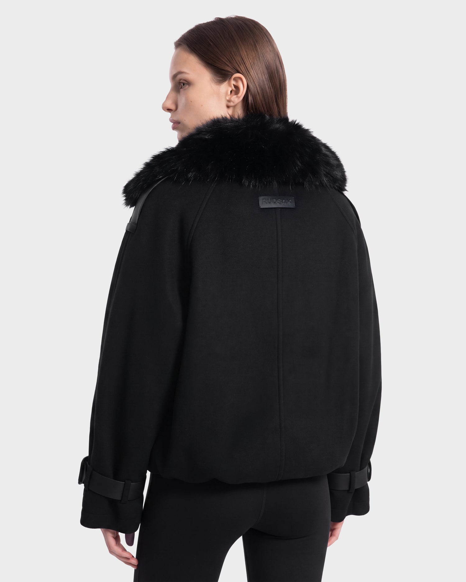 A woman with straight brown hair is seen from the back wearing the Rudsak MAEMI - 8126985 Black jacket, featuring a large faux fur collar. She stands against a plain light gray background, her head turned slightly to the side. This stylish piece is available for pre-order and is expected to ship by September 15th.