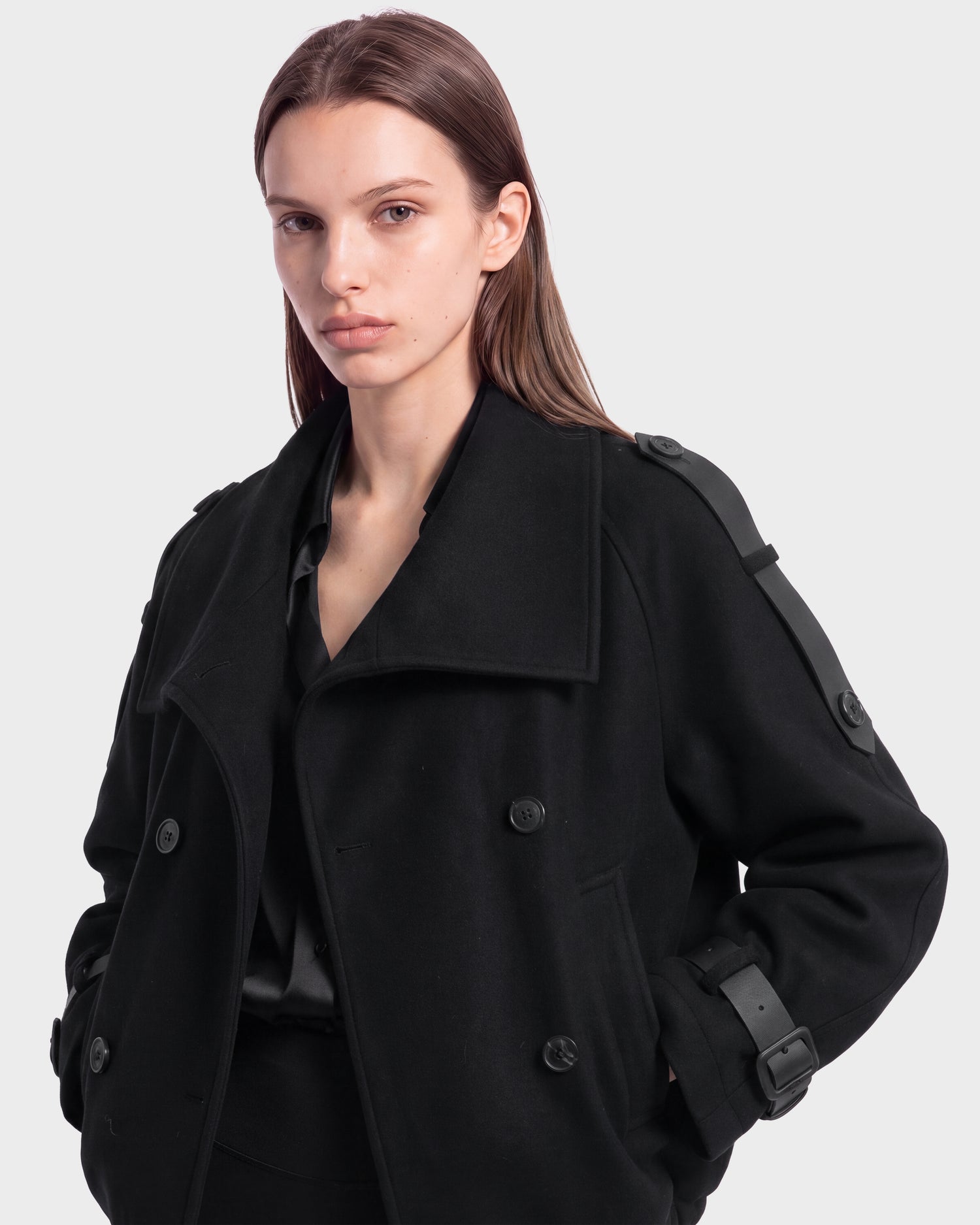 A person with long brown hair wears the MAEMI - 8126985 Black trench coat by Rudsak, featuring a double-breasted design and buckled cuffs, while standing against a plain light gray background. This stylish trench coat is available for pre-order now and is expected to ship by September 15th.