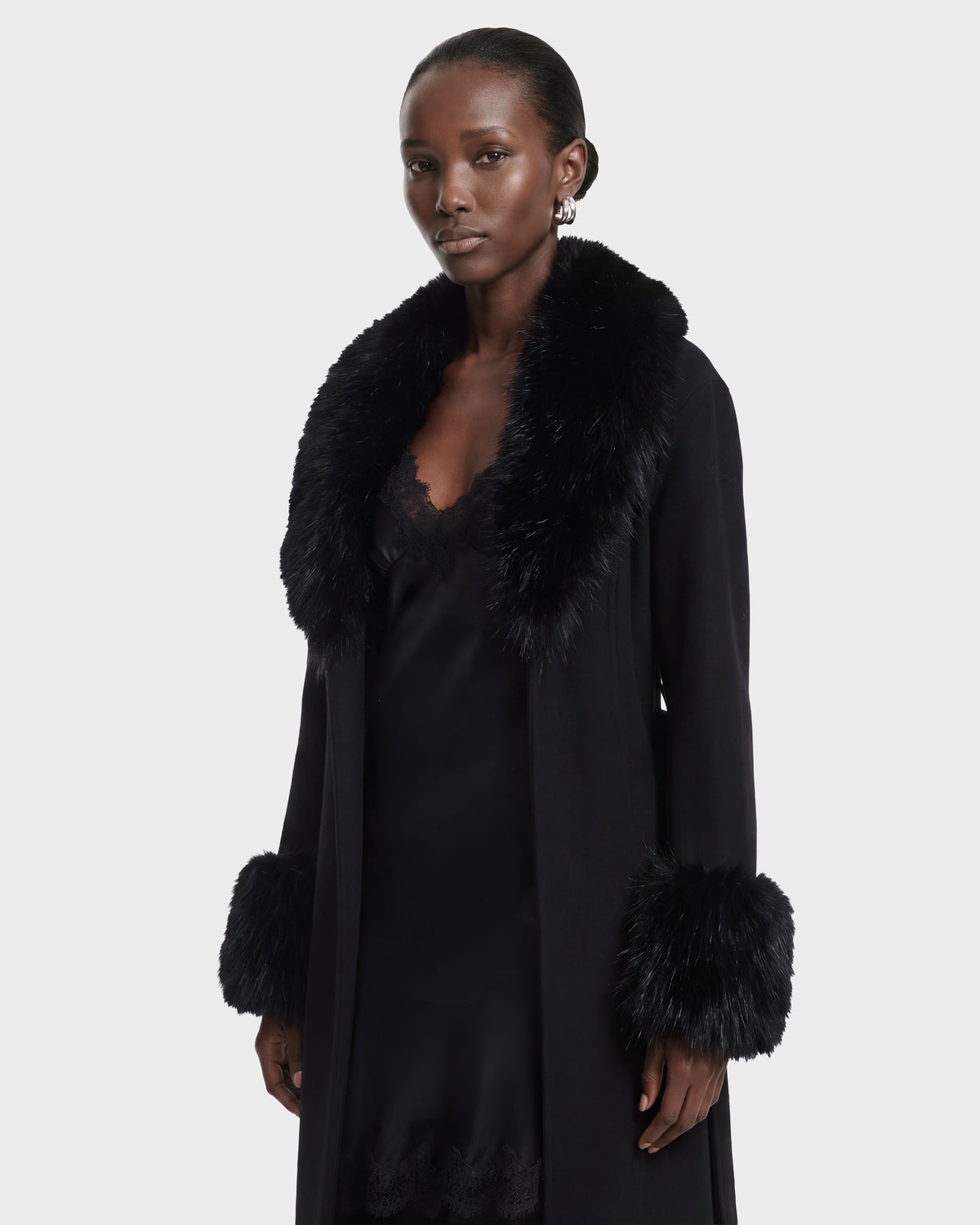 A person wearing the Rudsak MARILLA WL - 8126737 Black coat, featuring black fur trim on the collar and cuffs over a black dress, stands against a plain light background. This stylish look is available for pre-order and is expected to ship by September 15th.