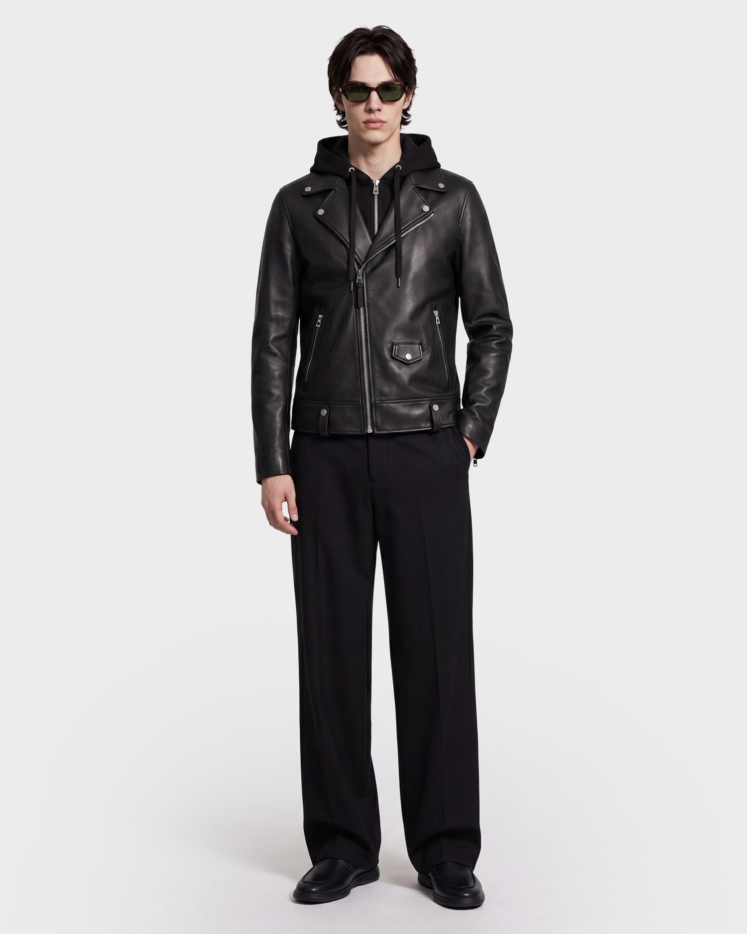 A person wearing sunglasses, the Rudsak MANUS - 6124600 Black men’s leather moto jacket with a removable hooded dickie layered over a hoodie, black wide-leg pants, and black shoes stands against a plain white background.