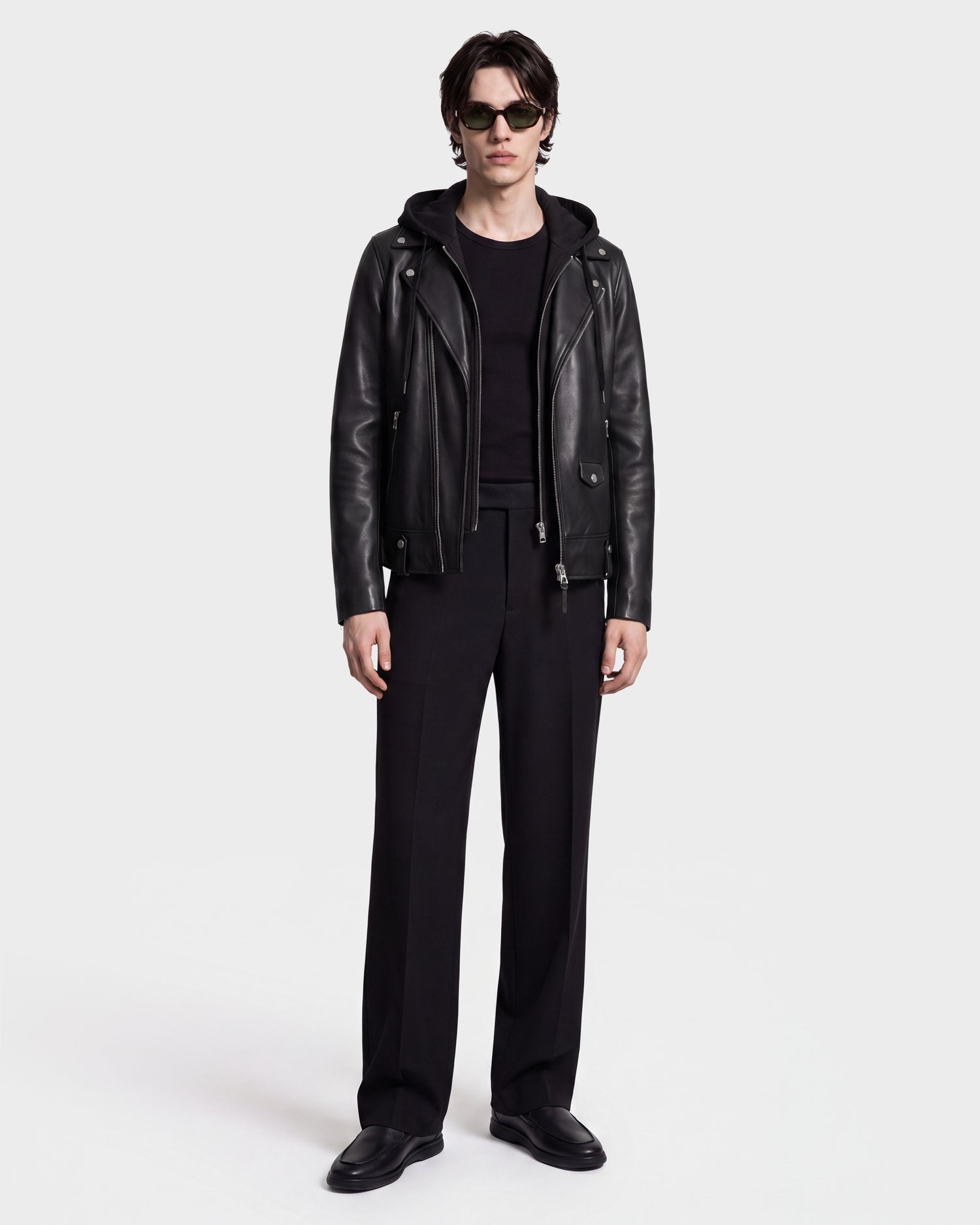 A person stands against a white background wearing sunglasses and the MANUS - 6124600 Black men’s leather moto jacket with a removable hooded dickie by Rudsak, layered over a black hoodie, along with a black shirt, black trousers, and black shoes.