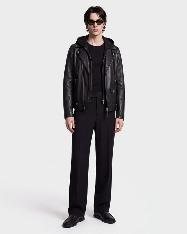 A person stands against a white background wearing sunglasses and the MANUS - 6124600 Black men’s leather moto jacket with a removable hooded dickie by Rudsak, layered over a black hoodie, along with a black shirt, black trousers, and black shoes.