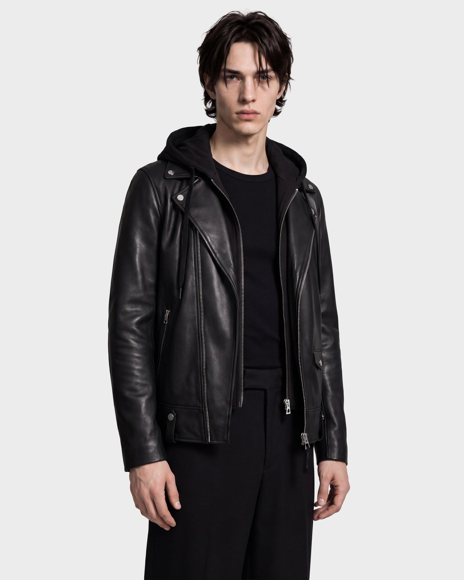 A person wearing the Rudsak MANUS - 6124600 Black men’s leather moto jacket with a removable hooded dickie over a black shirt and black pants stands facing forward against a plain light background.