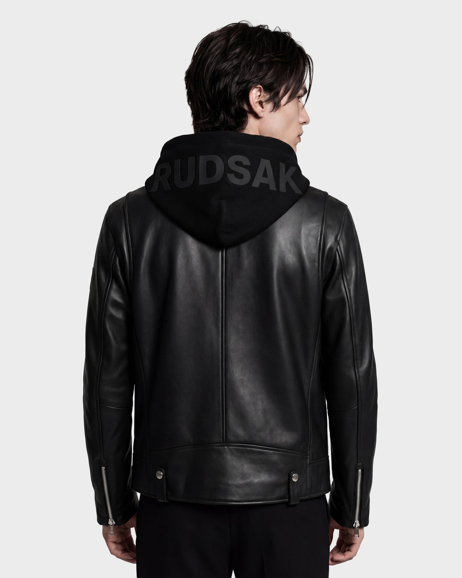 A person facing away, wearing the Rudsak MANUS - 6124600 Black men’s leather moto jacket layered over a black hooded sweatshirt with “RUDSAK” in bold lettering on the hood, showcasing a modern interpretation of the classic biker jacket style.