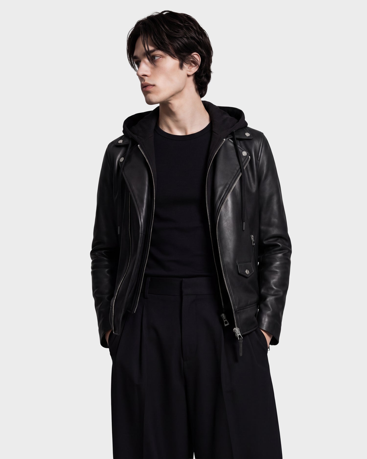 Wearing the Rudsak MANUS - 6124600 Black men’s leather moto jacket with a removable hooded dickie over a black shirt and loose black pants, a person with medium-length dark hair stands against a plain background, hands in pockets and looking to the side.