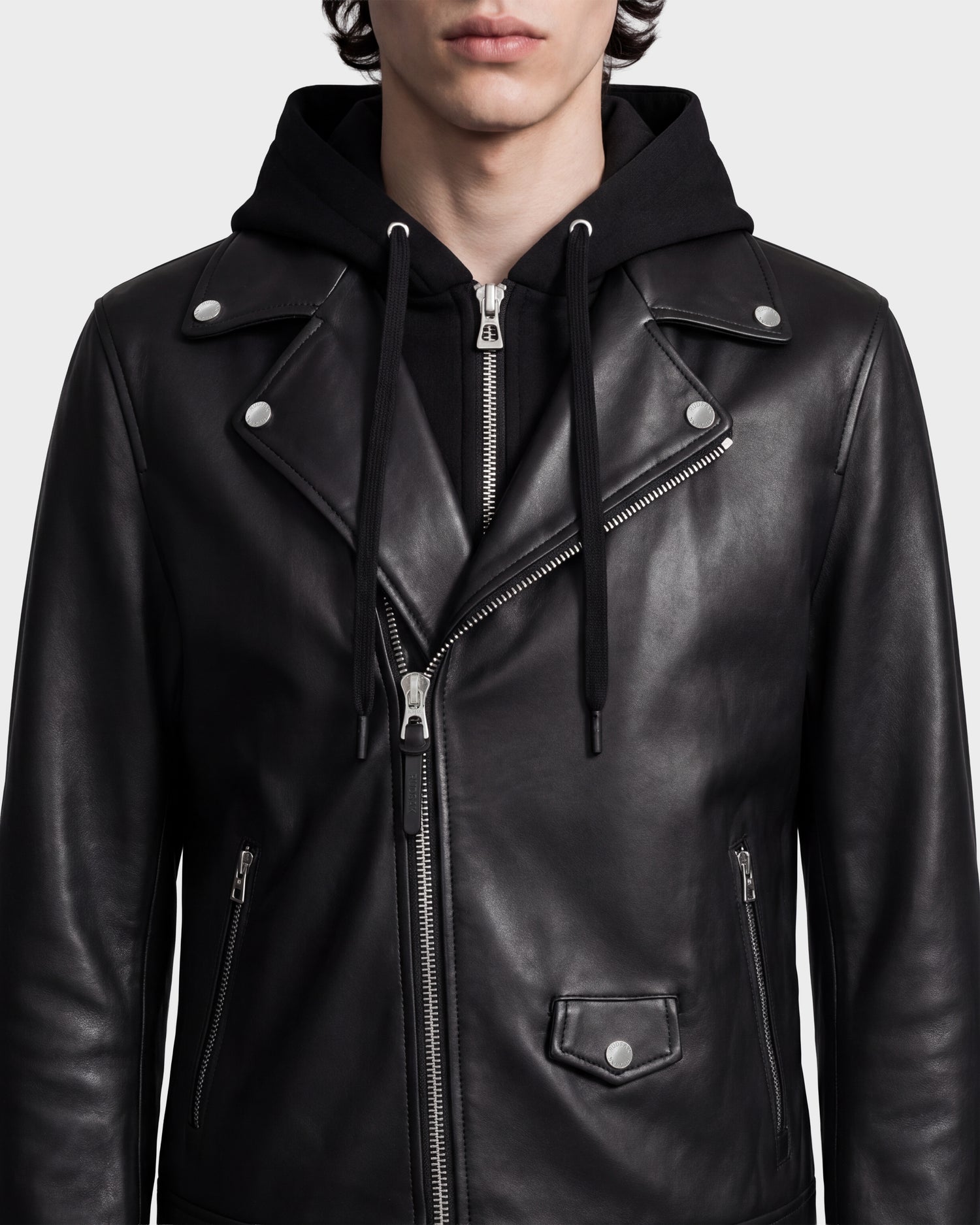 A person wearing the Rudsak MANUS - 6124600 Black men’s leather moto jacket with a removable hooded dickie over a black hoodie, shown from shoulders to mouth, against a plain light background.