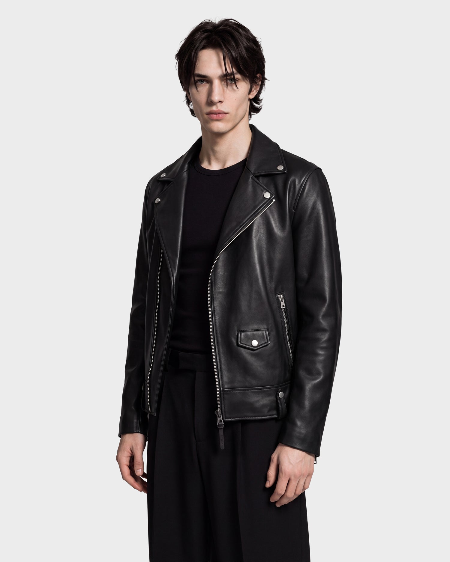 A person wearing the Rudsak MANUS - 6124600 Black men’s leather moto jacket with a removable hooded dickie over a black shirt and black pants stands against a plain light grey background.