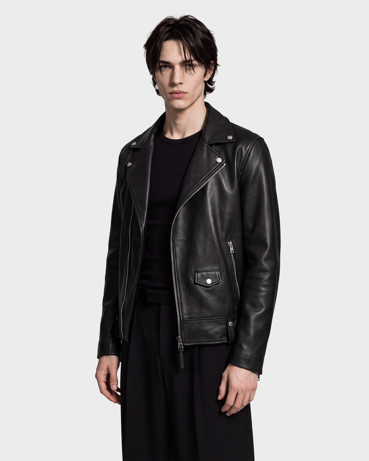 A person wearing the Rudsak MANUS - 6124600 Black men’s leather moto jacket with a removable hooded dickie over a black shirt and black pants stands against a plain light grey background.