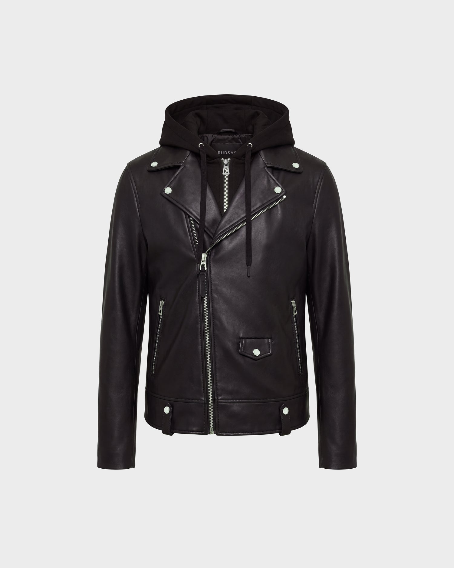 The MANUS - 6124600 Black men’s leather moto jacket by Rudsak features black leather construction with silver zippers and buttons, an asymmetrical front zip, two zippered pockets, a flap pocket on the front, and a removable hooded dickie for added versatility. The background is light gray.