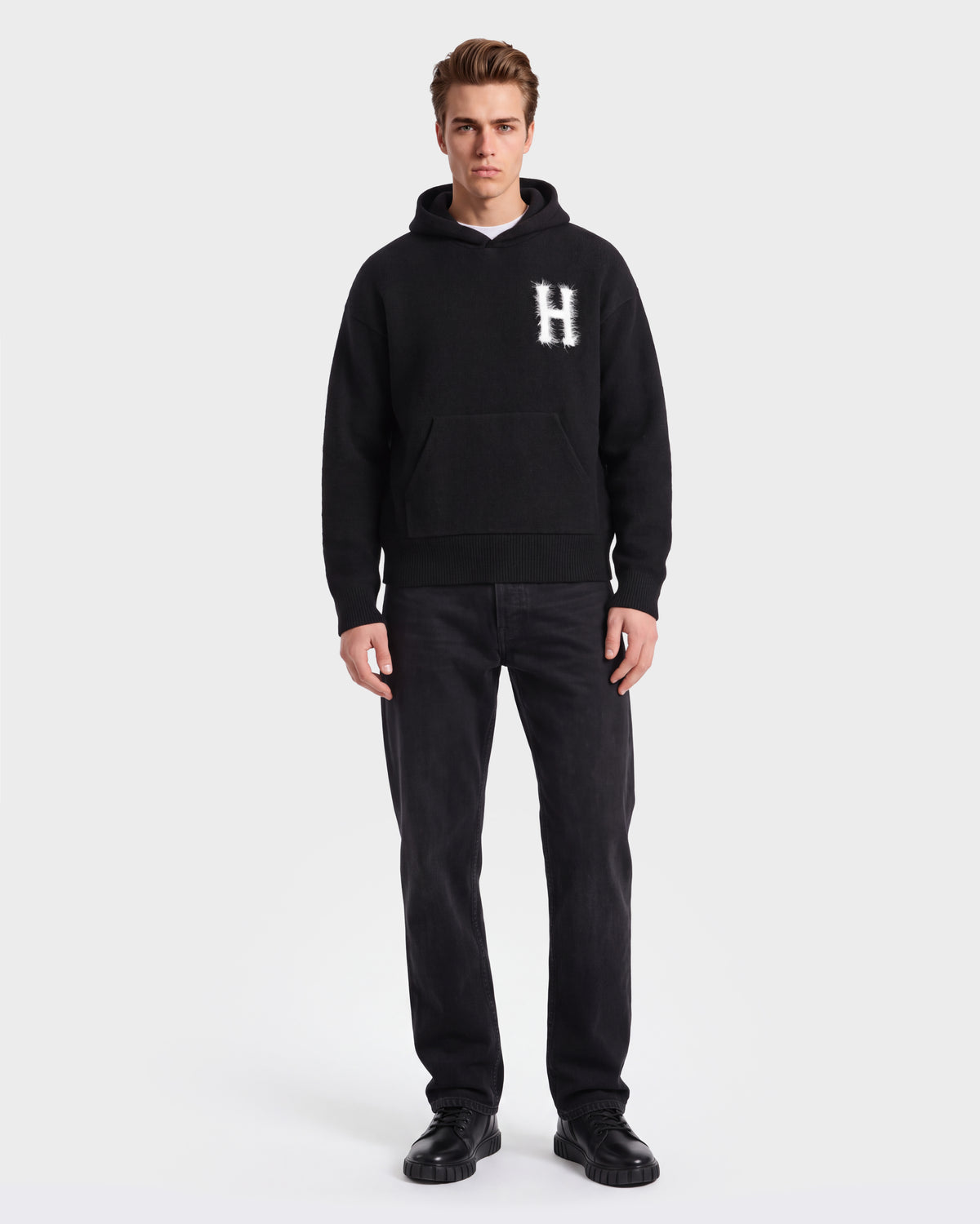 A person stands facing forward, wearing the Rudsak MARCUS HV - H525503 Black hoodie with a bold white H, paired with black pants and black shoes, set against a plain white background for a classic collegiate style look.