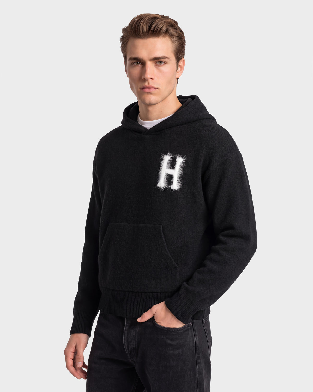 A person wearing the Rudsak MARCUS HV - H525503 Black hoodie, featuring a white “H” design on the chest, stands against a plain light background and looks at the camera with a neutral expression, effortlessly showcasing collegiate style.