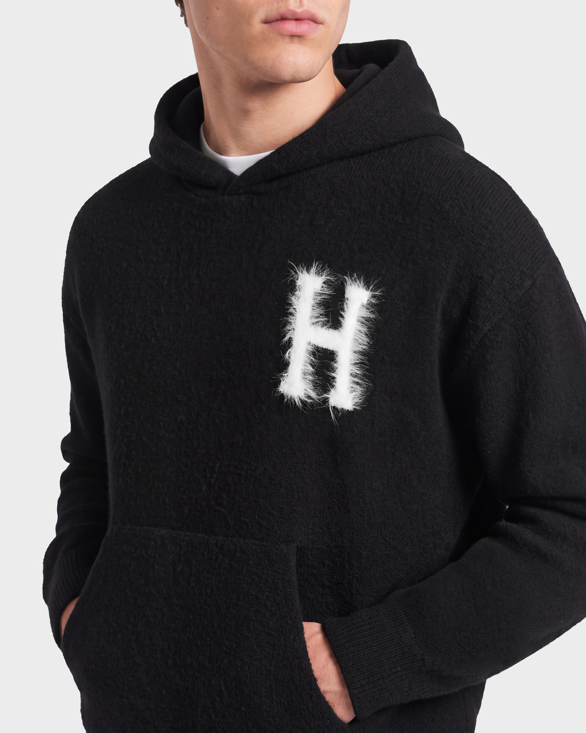 Wearing the Rudsak MARCUS HV - H525503 Black hoodie, featuring a furry white letter H on the chest, this individual exudes an effortlessly cool collegiate style. With one hand tucked into the front pocket and their face partially out of frame, their look is casual yet standout.