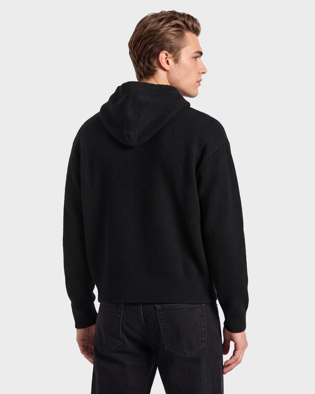 A person stands facing away, wearing the Rudsak MARCUS HV - H525503 Black hoodie and black jeans against a plain light gray background.