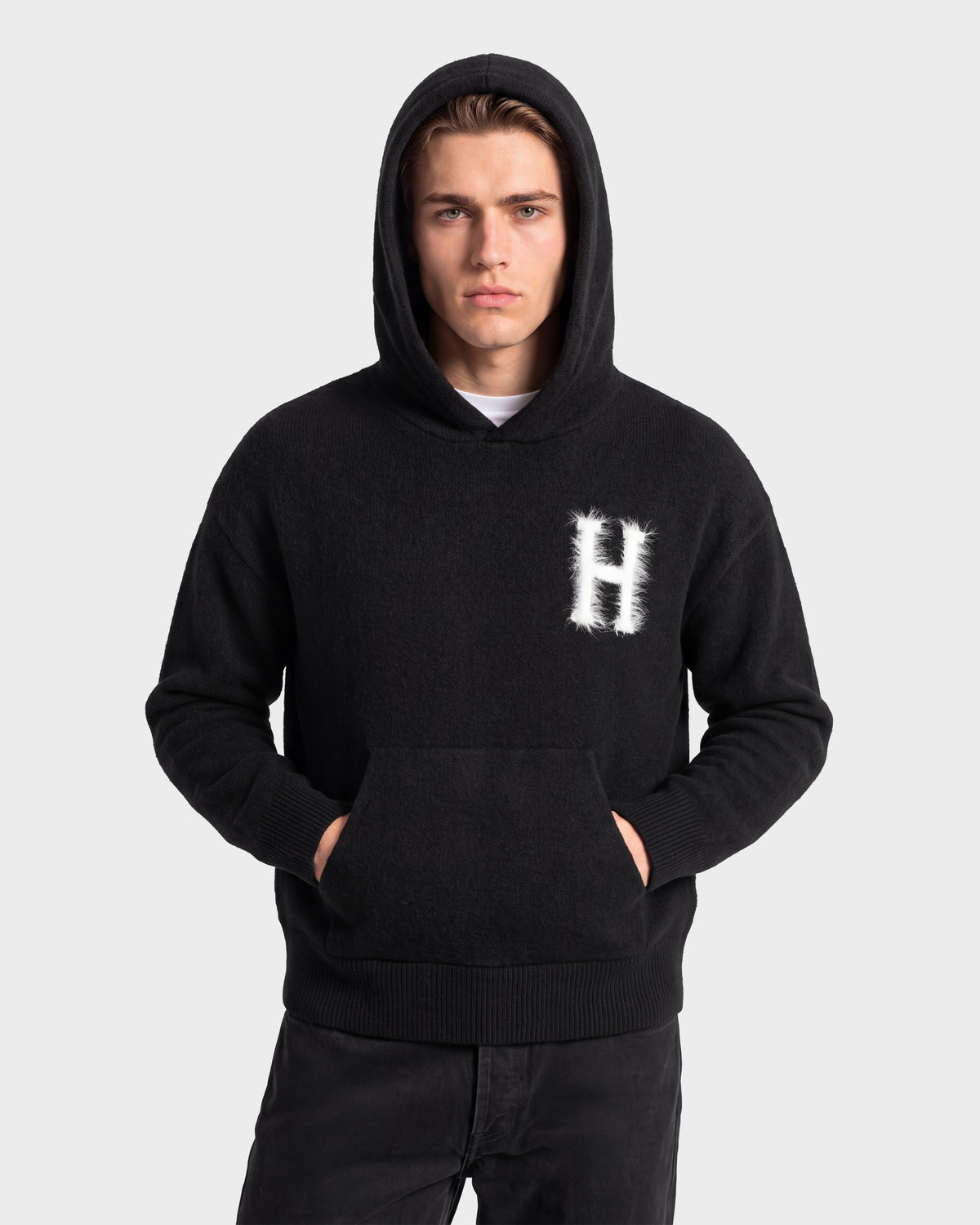 A person wearing the Rudsak MARCUS HV - H525503 Black hoodie, featuring a prominent H on the chest, stands with hands in the front pocket facing forward against a plain light gray background, showcasing a timeless collegiate style.