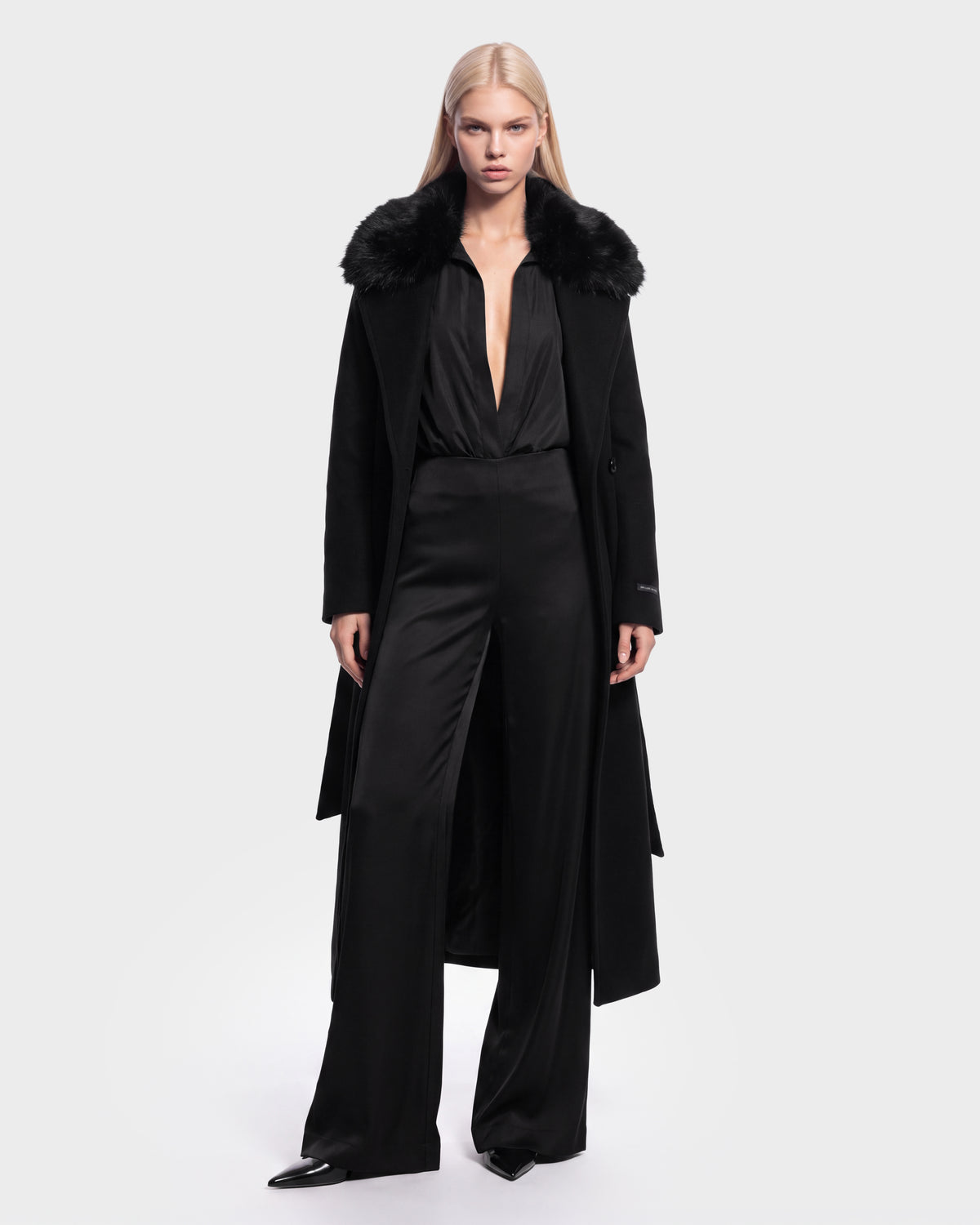 A woman with long blonde hair wears the Rudsak MEL - 8126525 Black coat with a fur collar over a deep V-neck black jumpsuit and pointed black shoes, standing against a plain white background. This stylish look is available for pre-order and is expected to ship by September 15th.