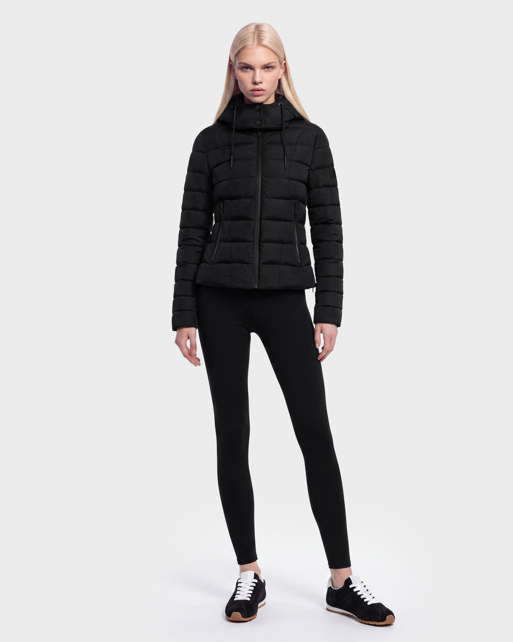 A woman with long blonde hair wears the Rudsak MELISSA - 8125054 Black women’s puffer jacket, filled with RDS certified down, paired with black leggings and black sneakers, standing against a plain white background.