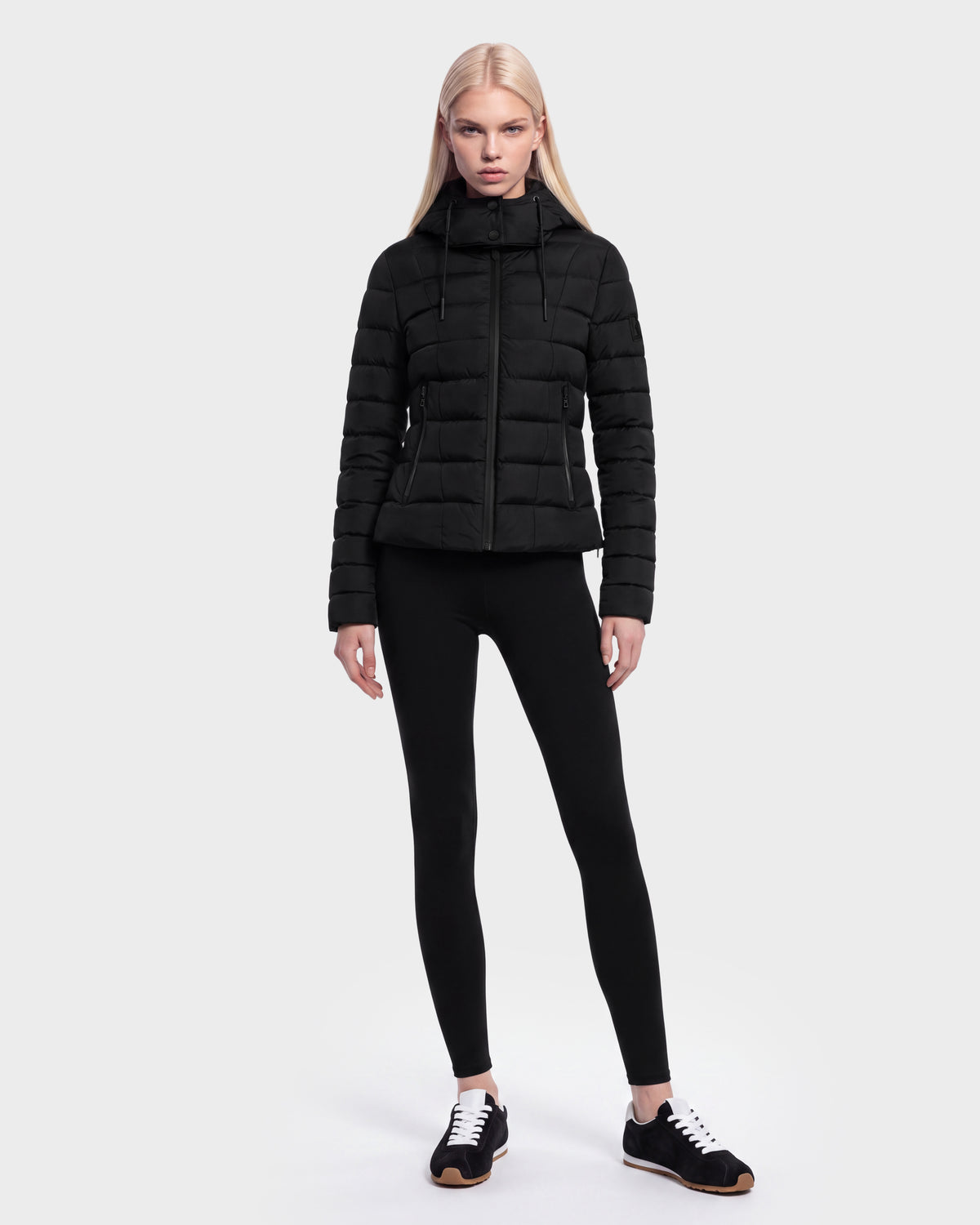A woman with long blonde hair wears the Rudsak MELISSA - 8125054 Black women’s puffer jacket, filled with RDS certified down, paired with black leggings and black sneakers, standing against a plain white background.