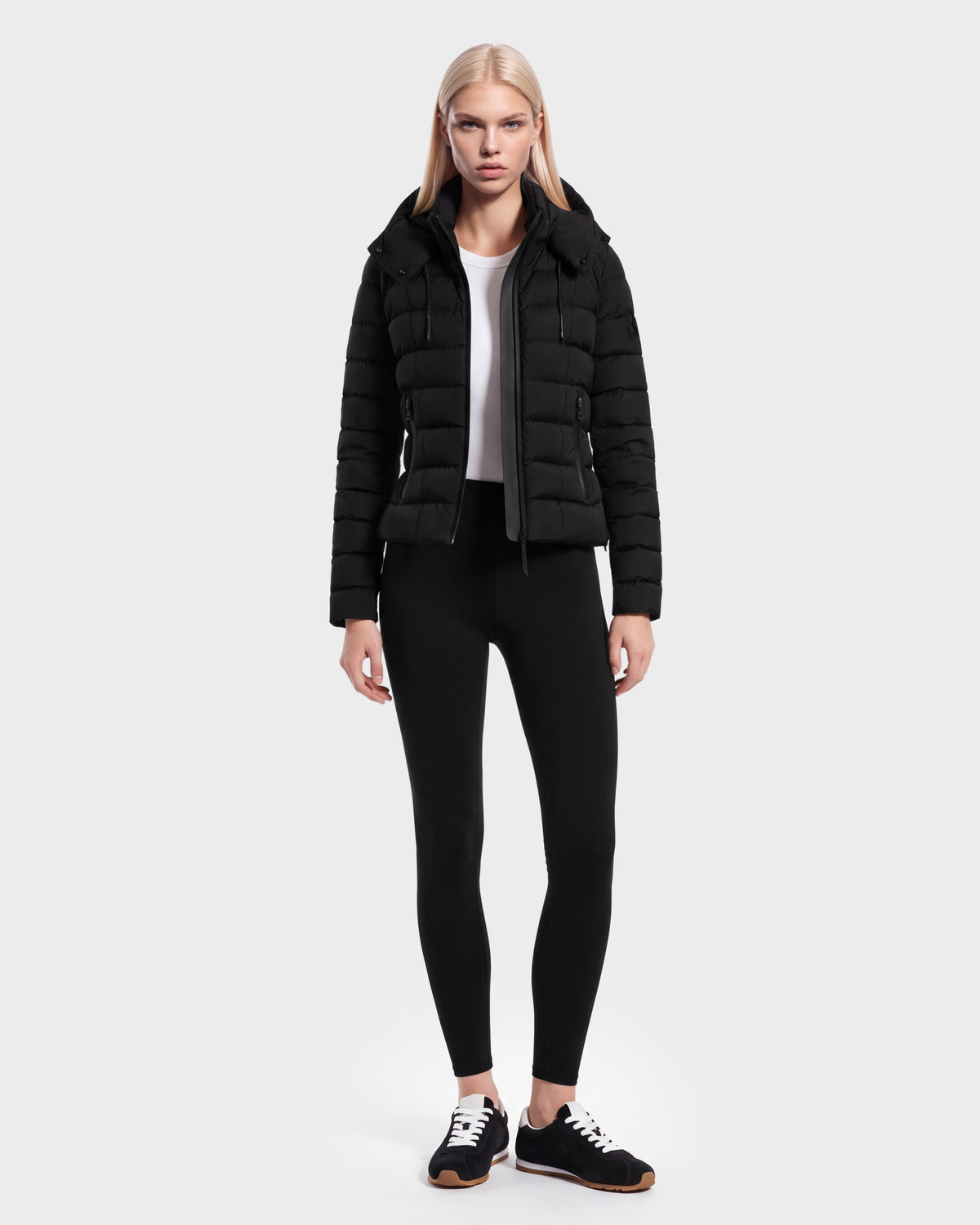 A woman stands facing forward against a plain light background, wearing the Rudsak MELISSA - 8125054 Black women’s puffer jacket crafted with RDS certified down, paired with a white shirt, black leggings, and black sneakers with white soles.