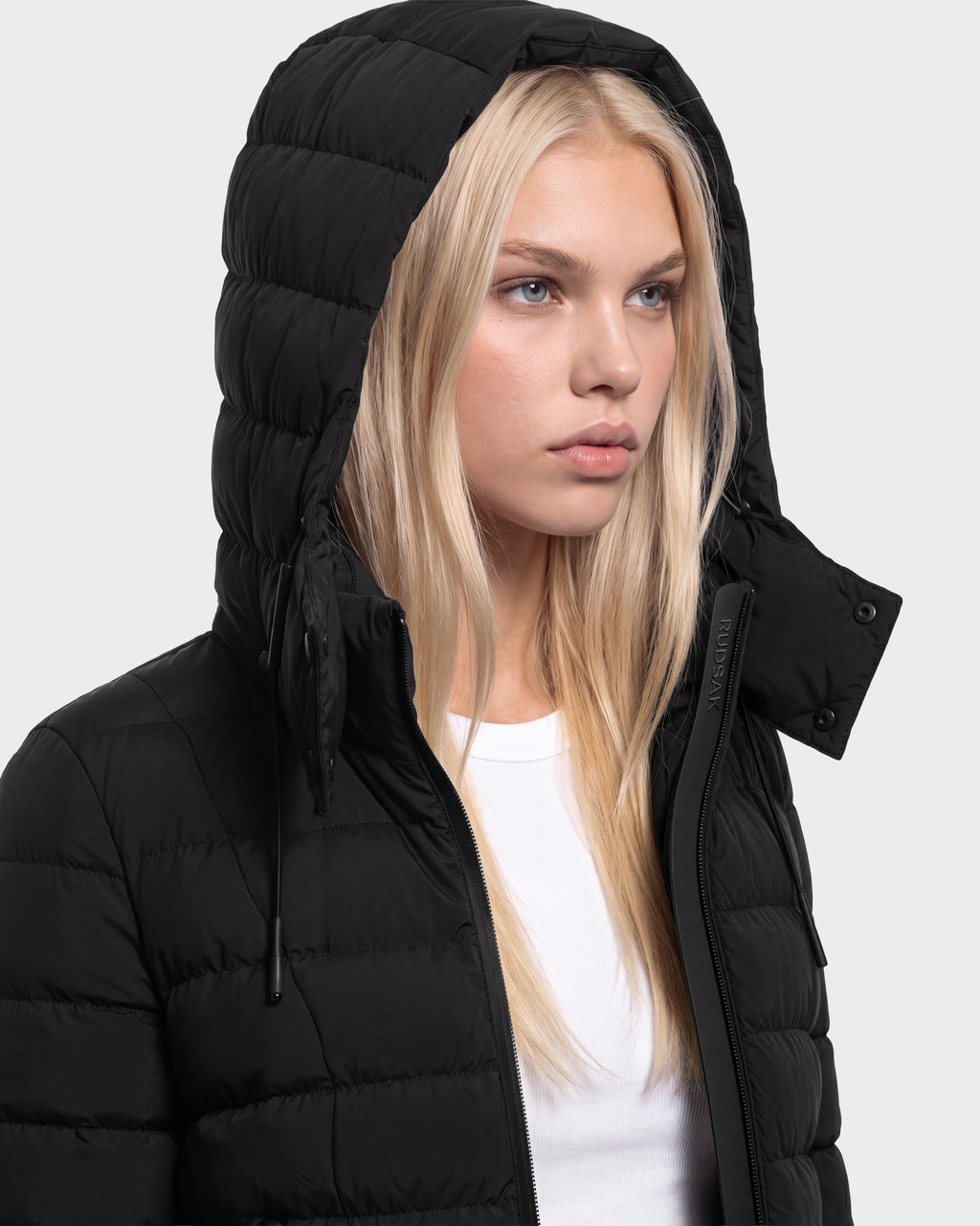 A person with long blonde hair is wearing the Rudsak MELISSA - 8125054 Black women’s puffer jacket with the hood up over a white shirt, looking slightly to the side against a plain light background.