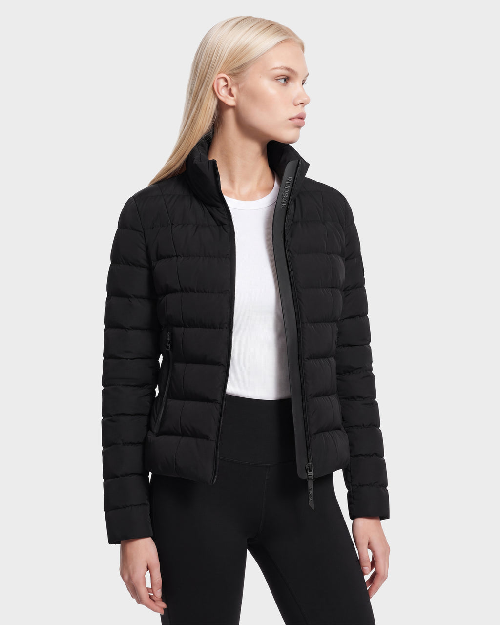 A person with straight blond hair wears the MELISSA - 8125054 Black women’s puffer jacket by Rudsak over a white shirt and black pants, standing against a plain light gray background, looking to the left.