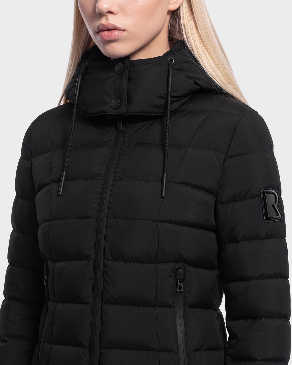 A woman with long blonde hair wears the MELISSA - 8125054 Black women’s puffer jacket from Rudsak. Made with RDS certified down, this black jacket features a high collar, zipper closure, and an R patch on the sleeve. Only the lower half of her face is visible.