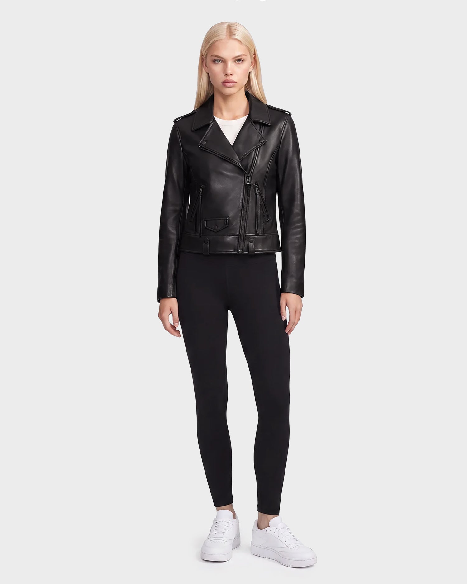 A woman with long blonde hair wears the Rudsak MERGO - 8122009 BLACK lambskin leather biker jacket, paired with black leggings and white sneakers. She stands facing forward against a plain light gray background.