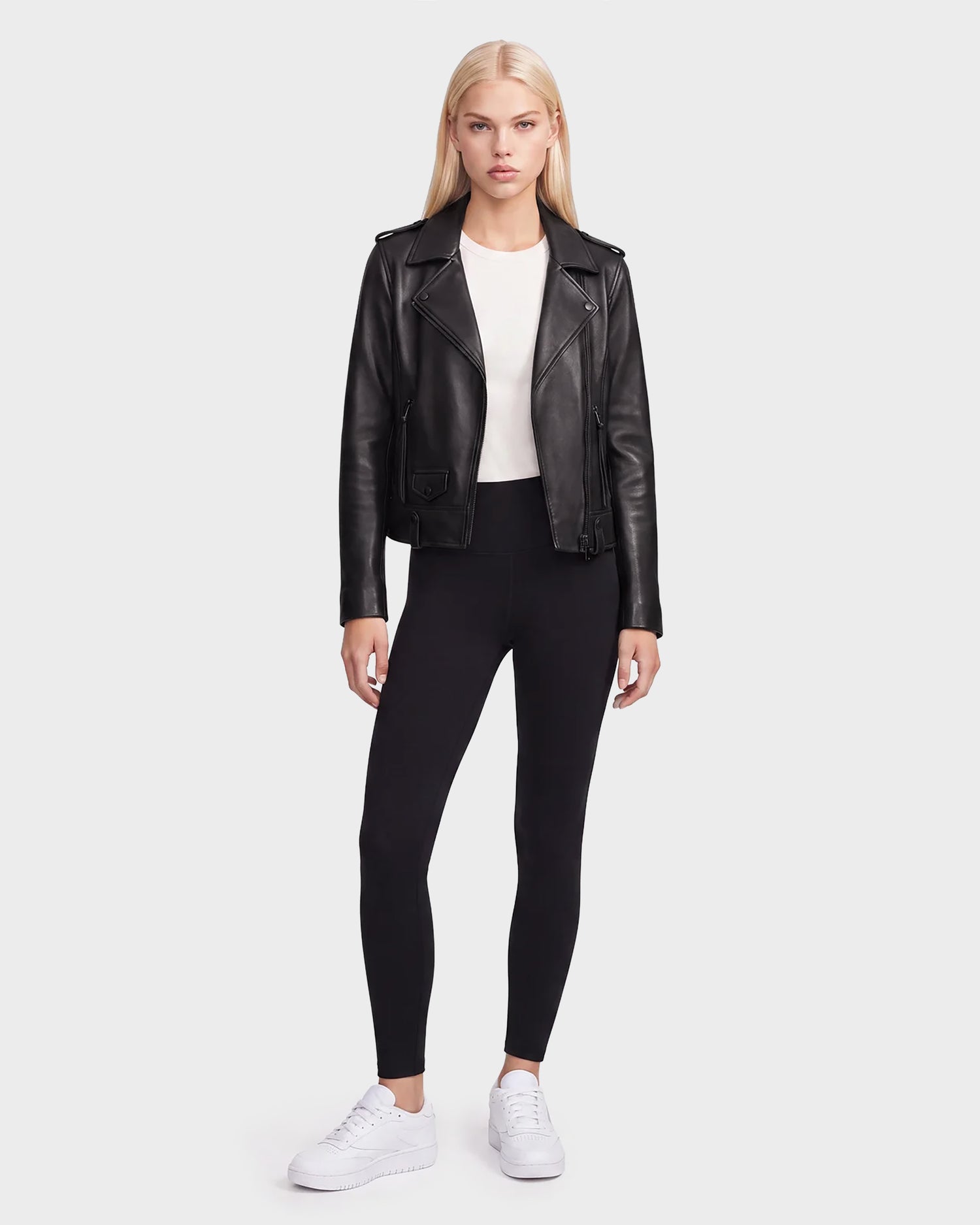 A woman stands facing forward against a plain light background, wearing the Rudsak MERGO - 8122009 BLACK lambskin leather biker jacket, styled with a white shirt, black leggings, and white sneakers.