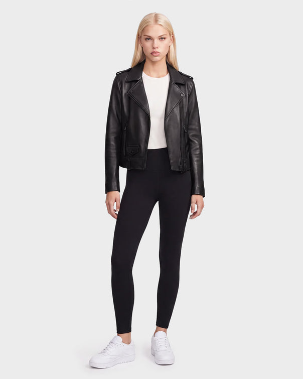A woman stands facing forward against a plain light background, wearing the Rudsak MERGO - 8122009 BLACK lambskin leather biker jacket, styled with a white shirt, black leggings, and white sneakers.