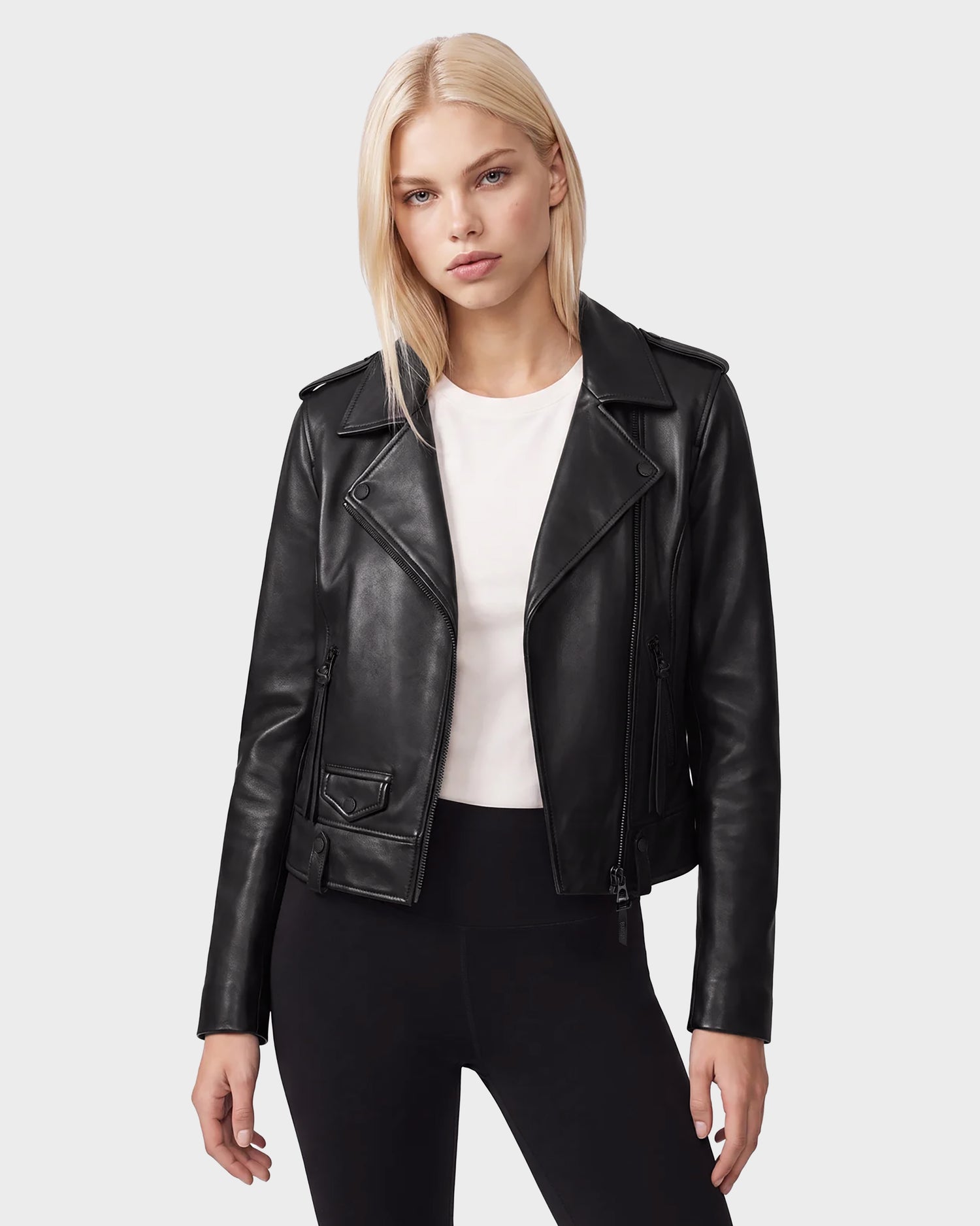 A woman with straight blonde hair wears the Rudsak MERGO - 8122009 BLACK lambskin leather biker jacket over a white t-shirt and black pants, standing against a plain light gray background.