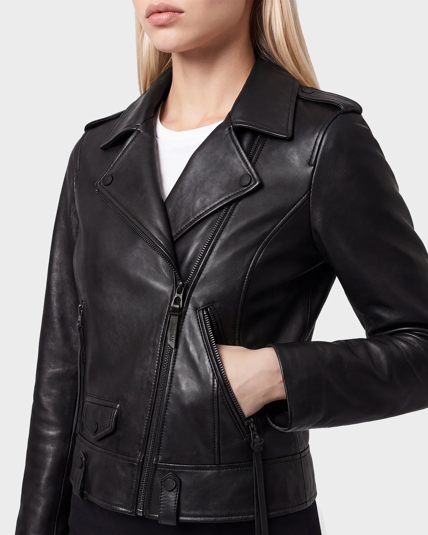 A woman with long blonde hair wears the Rudsak MERGO - 8122009 BLACK lambskin leather biker jacket over a white top, with one hand in the jacket pocket, shown from the shoulders to the waist against a light background.