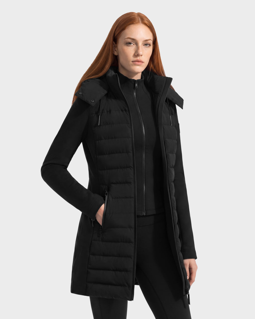 A woman wears a black, knee-length, hooded quilted down jacket from the Rudsak MERINA - 8125056 Black collection over a matching long-sleeved zip-up vest to create a versatile transitional look.