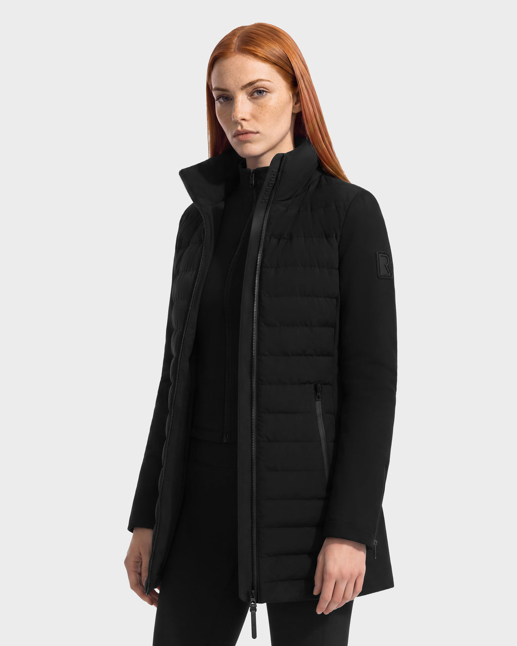 A model wearing the Rudsak MERINA - 8125056 Black, a knee-length, quilted down jacket with a high collar and zippered pockets.