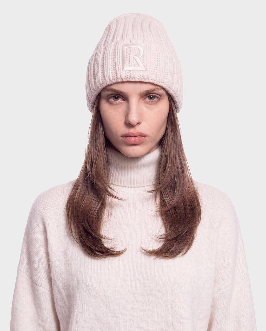 A person wearing a MISSA - 8426512 Vanilla cream-colored ribbed knit beanie with a letter R emblem on the front and a matching turtleneck sweater.