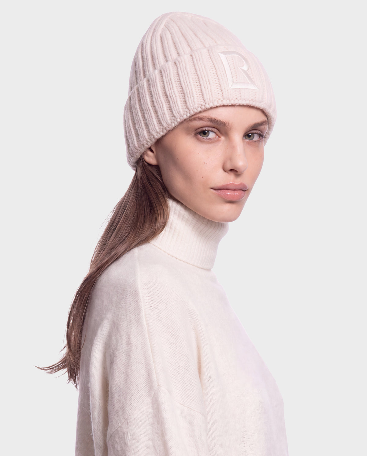 A person wearing a vanilla-colored MISSA - 8426512 beanie with a square logo patch on the ribbed cuff and a matching high-neck sweater.