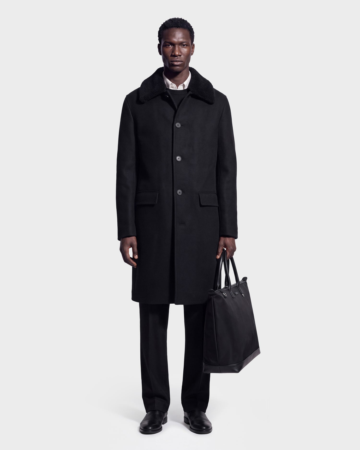 A man stands against a plain light gray background, looking straight at the camera, wearing the Rudsak NICO - 6126717 Black men’s long wool coat with a removable faux-shearling collar, paired with black pants, black shoes, and holding a black tote bag.